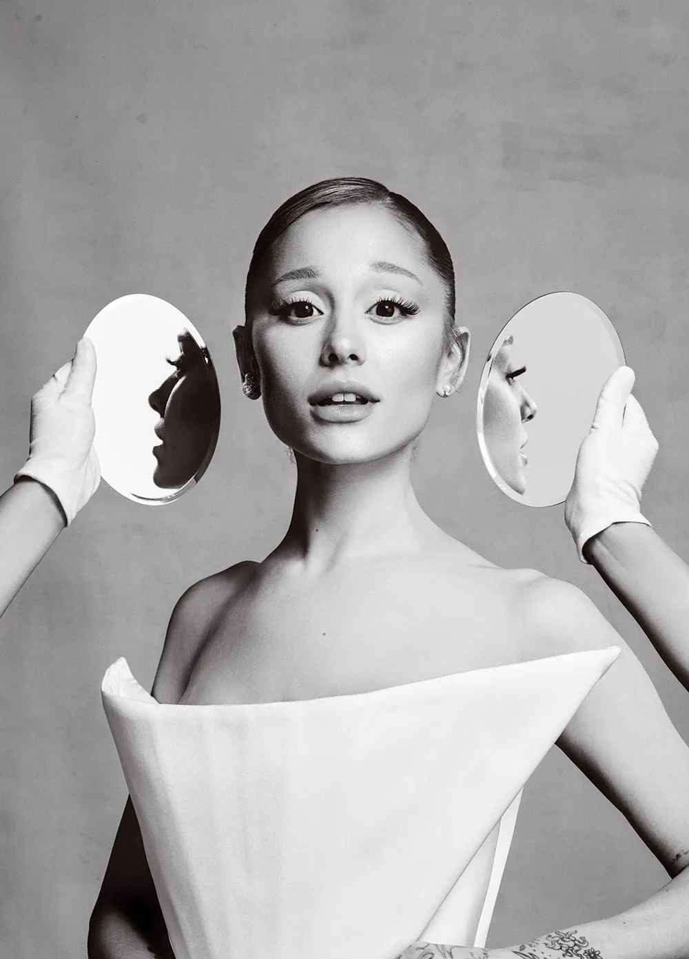 Close-up of Ariana Grande with two round mirrors held beside her face, showing profile reflections, black and white.