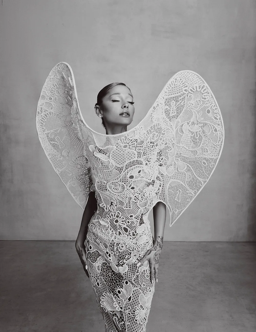 Ariana Grande in black and white, wearing an avant-garde lace gown with winged details and eyes closed.