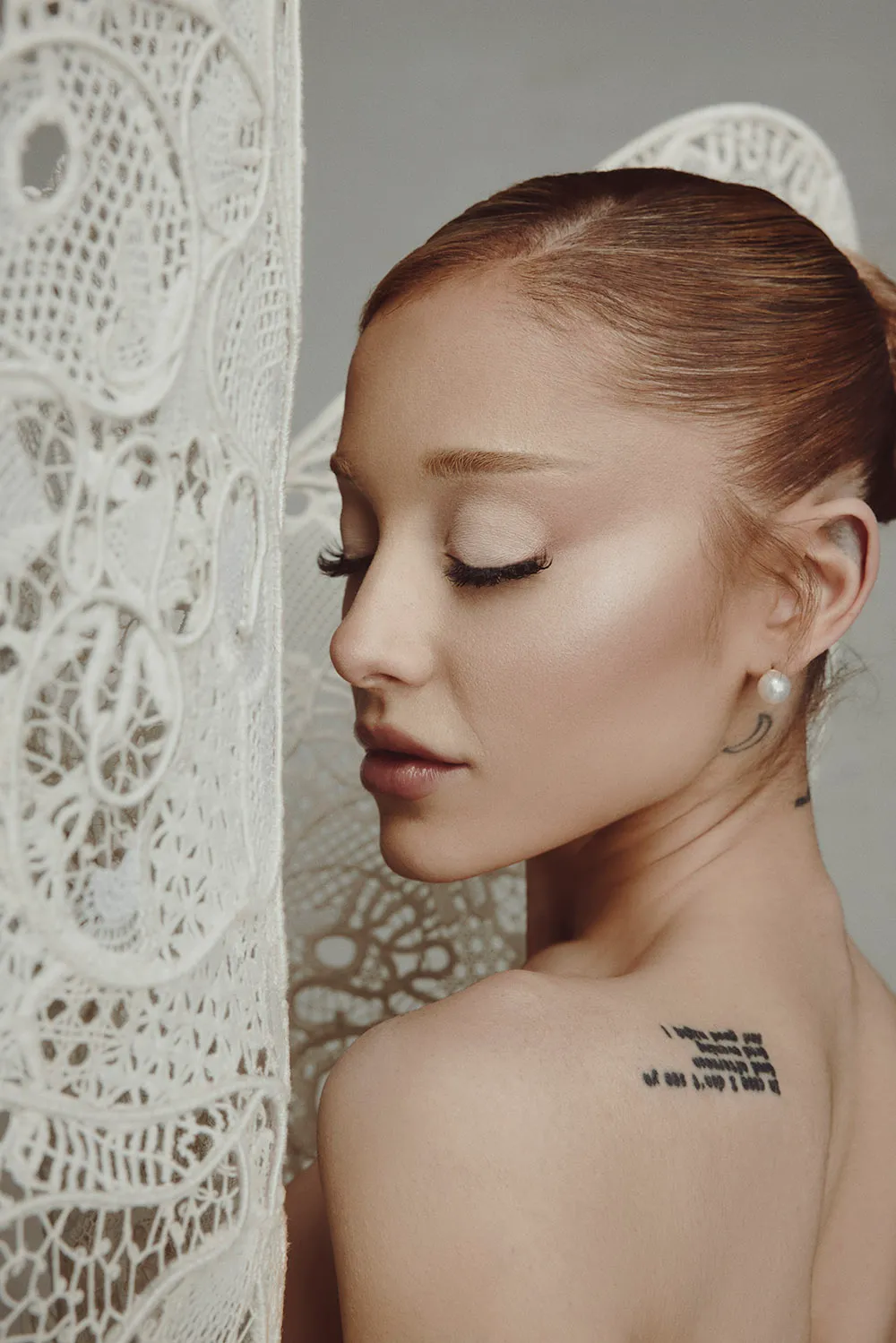 Close-up profile of Ariana Grande with her eyes closed, showing tattoo and intricate lace fabric beside her.