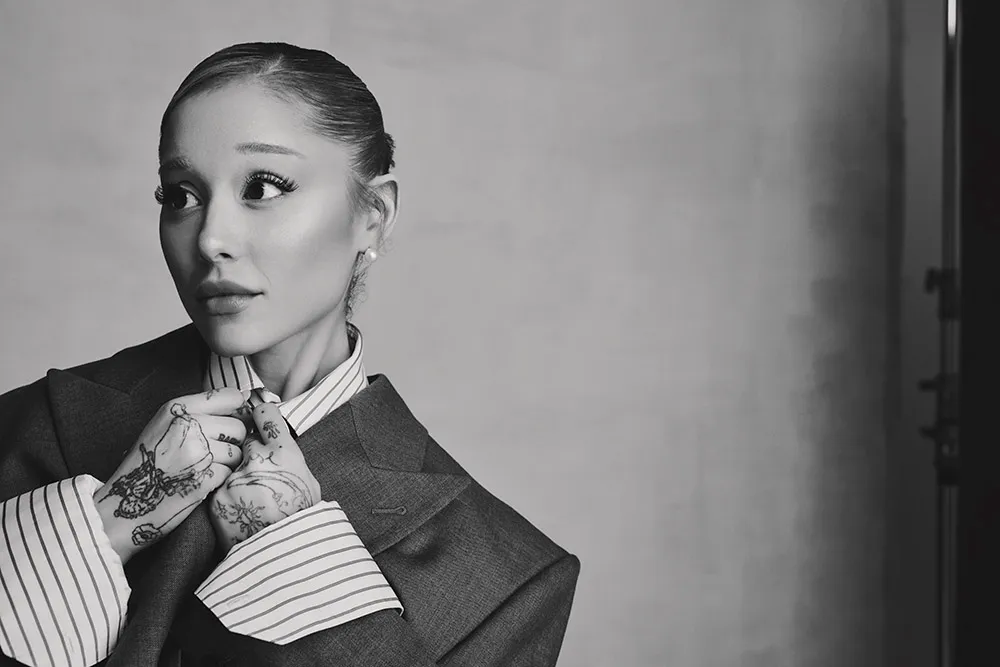 Black and white close-up of Ariana Grande adjusting her oversized suit collar with tattooed hands and pearl earring.