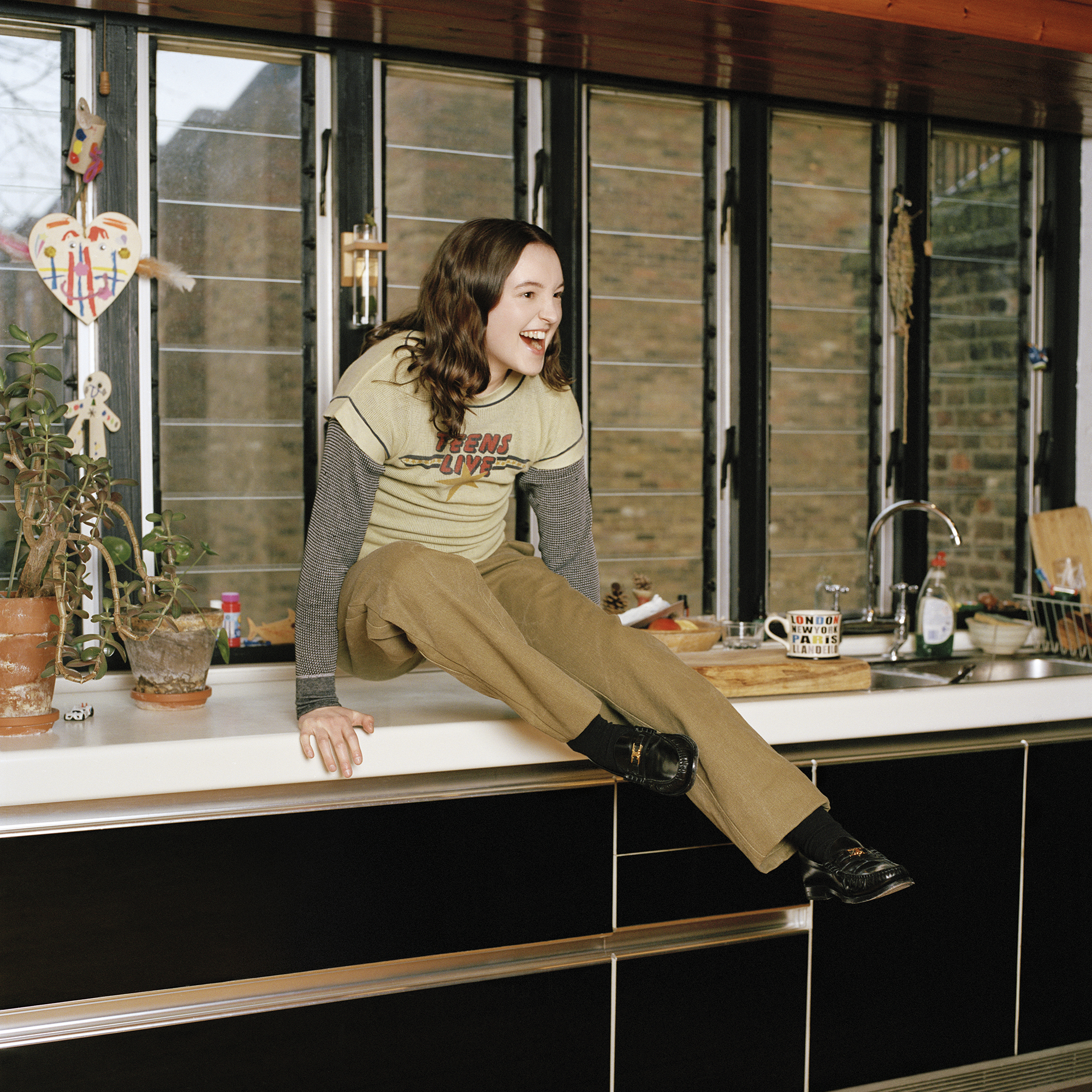 Bella Ramsey laughs while sitting on a kitchen counter in layered vintage-style outfit for Interview Magazine, May 2025