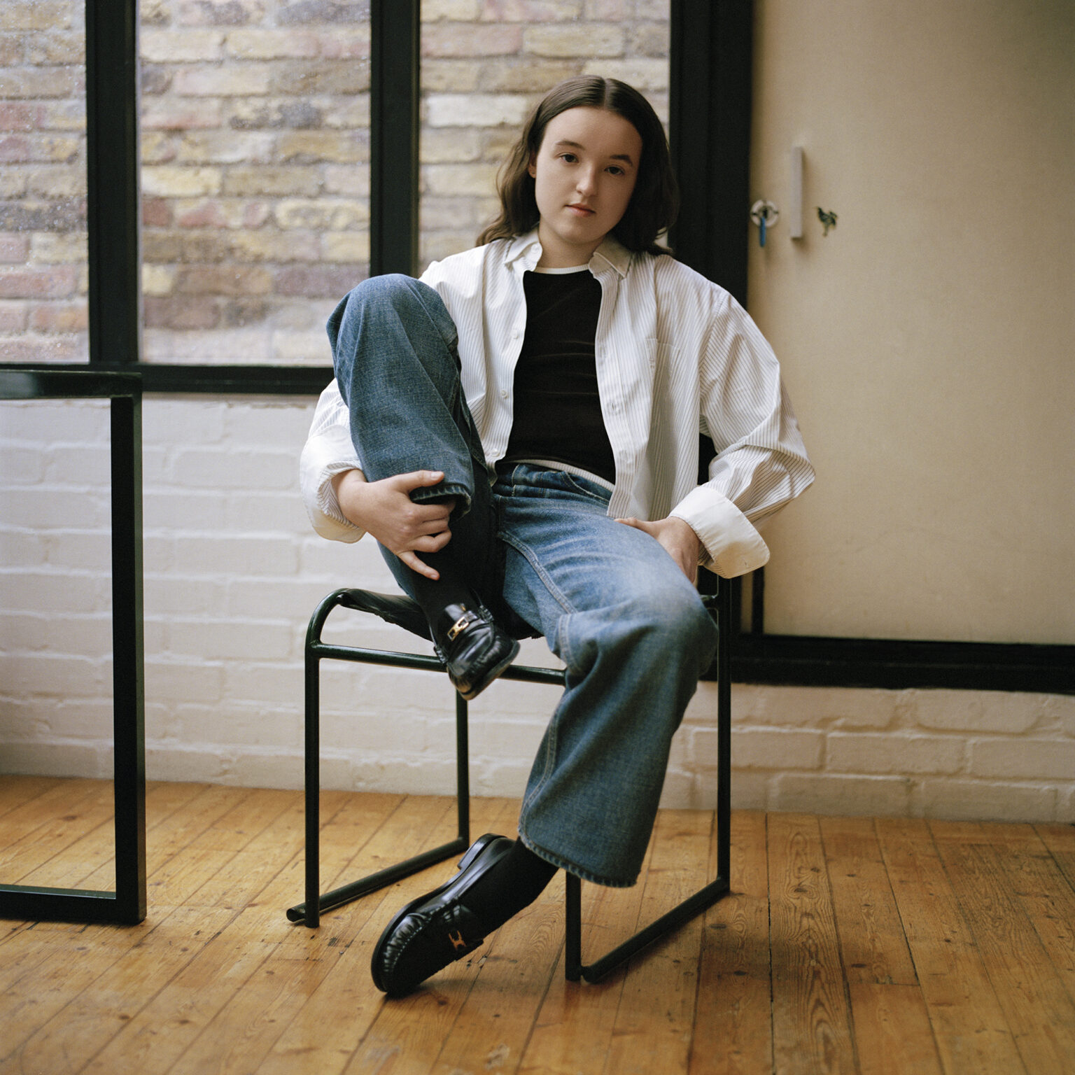 Bella Ramsey sits in a modern chair wearing striped shirt and jeans during her Interview Magazine editorial, May 2025