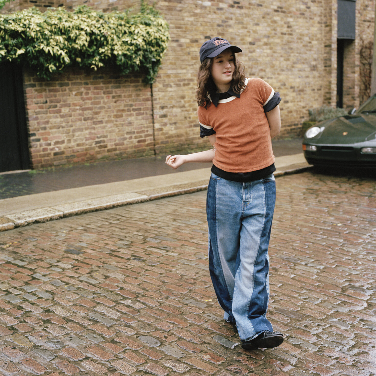 Bella Ramsey walks on cobblestone street in layered casualwear and cap for Interview Magazine shoot, May 2025