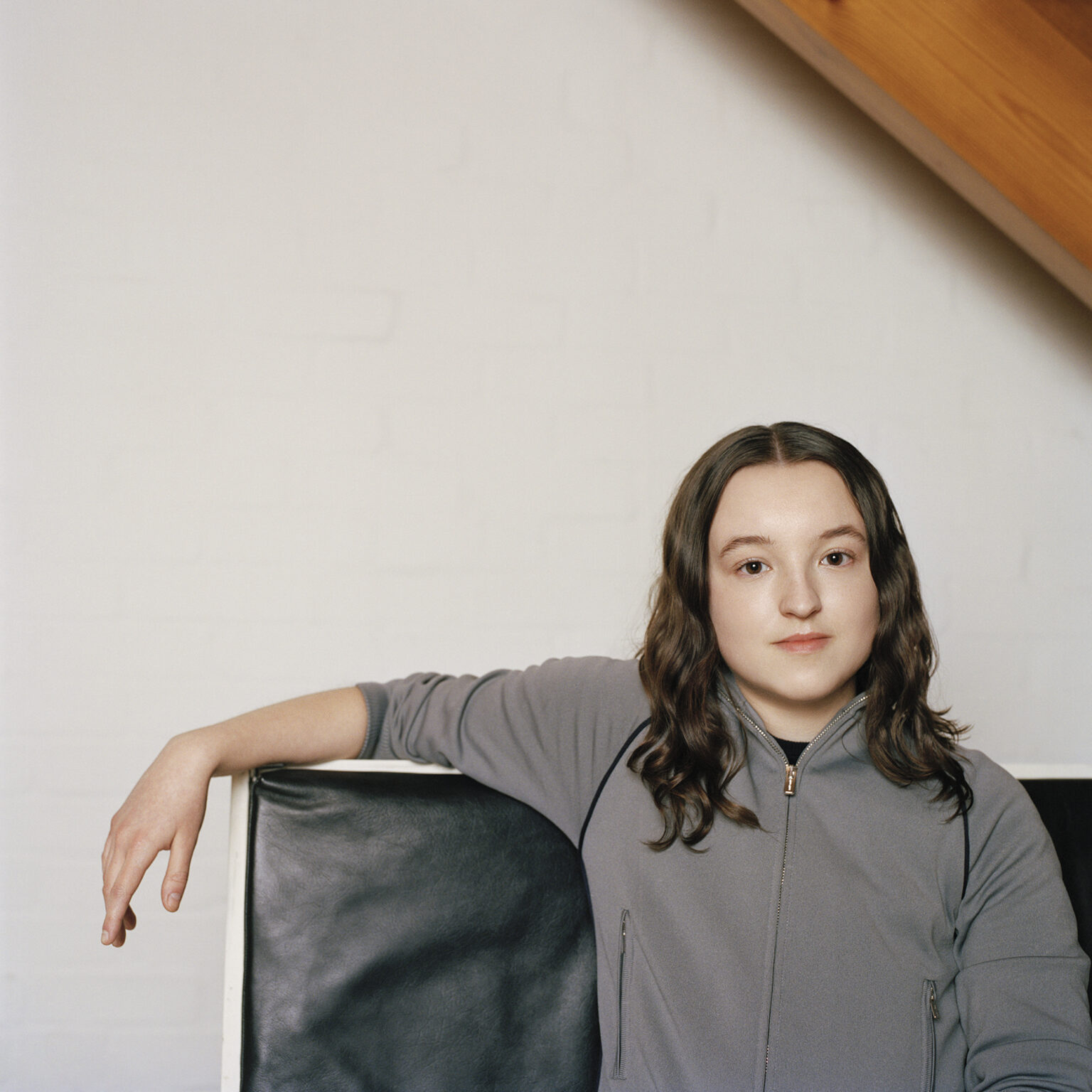 Bella Ramsey leans back on a black chair in a sporty zip-up jacket in indoor Interview Magazine portrait, May 2025