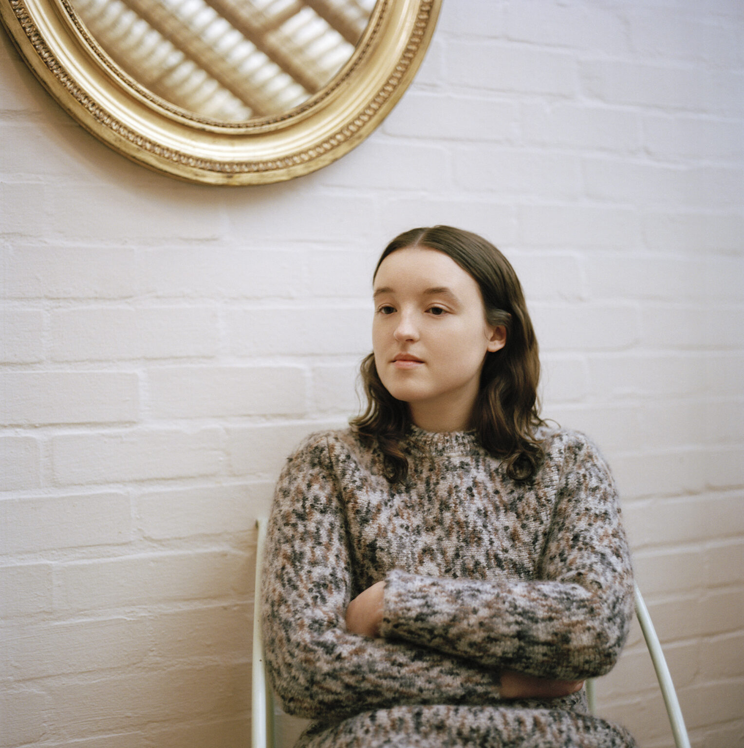 Bella Ramsey sits with arms crossed in cozy speckled sweater beneath a gold mirror for Interview Magazine, May 2025