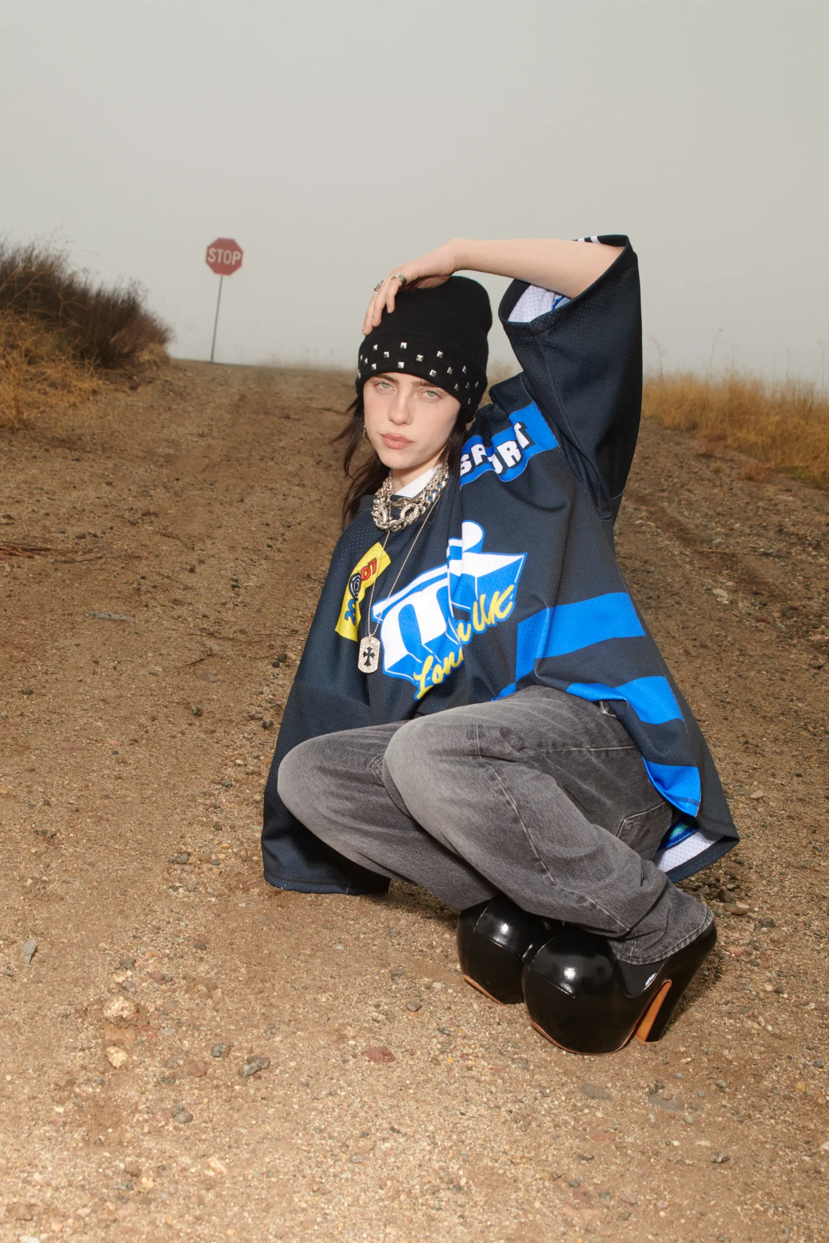 Billie Eilish crouches on a foggy dirt road in a beanie and oversized graphic jersey for British Vogue May 2025, styled with chunky heels and silver chains.
