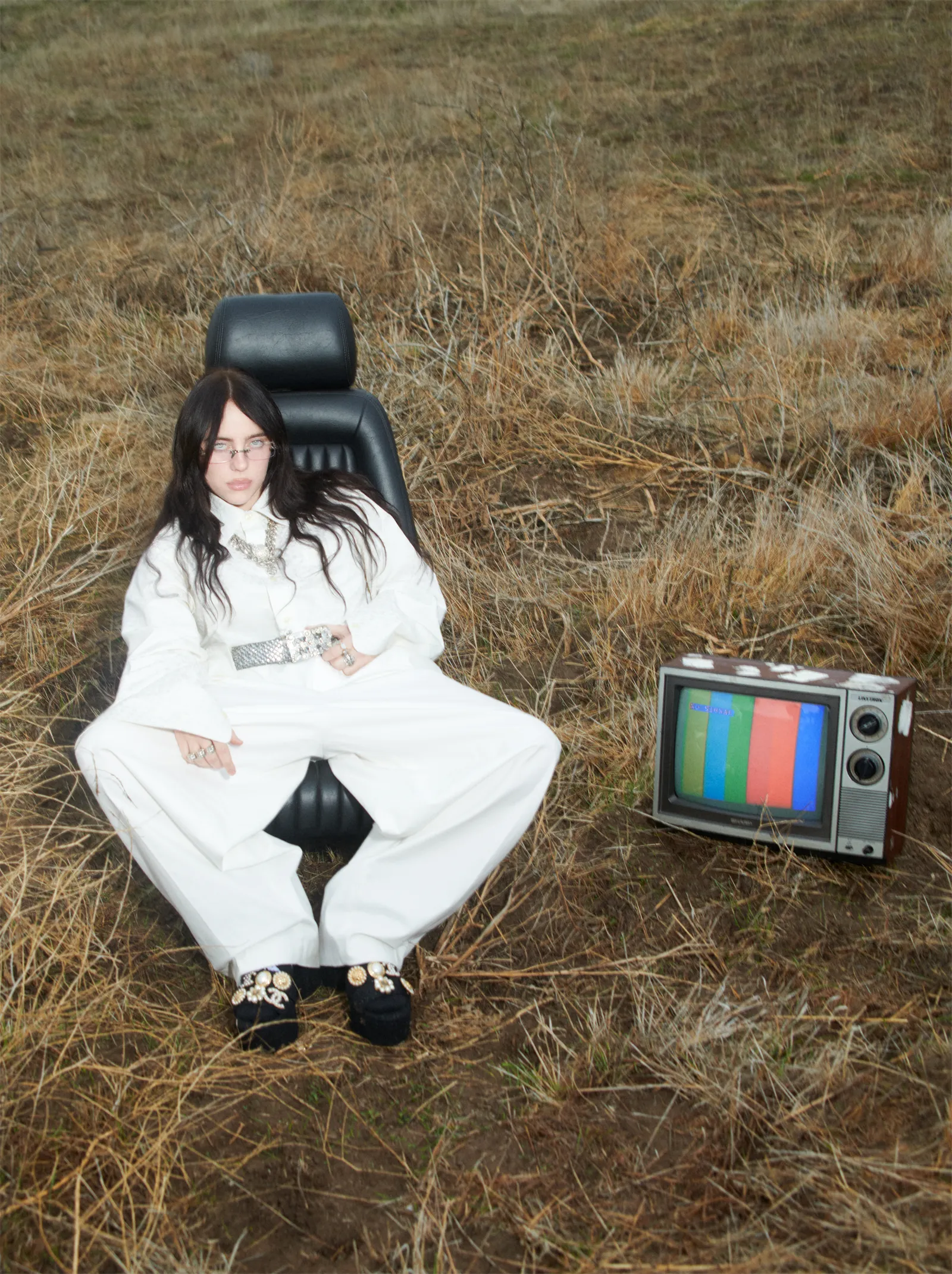 Billie Eilish lounges in an open field wearing an all-white oversized outfit and jeweled platform shoes beside a vintage TV for British Vogue May 2025.