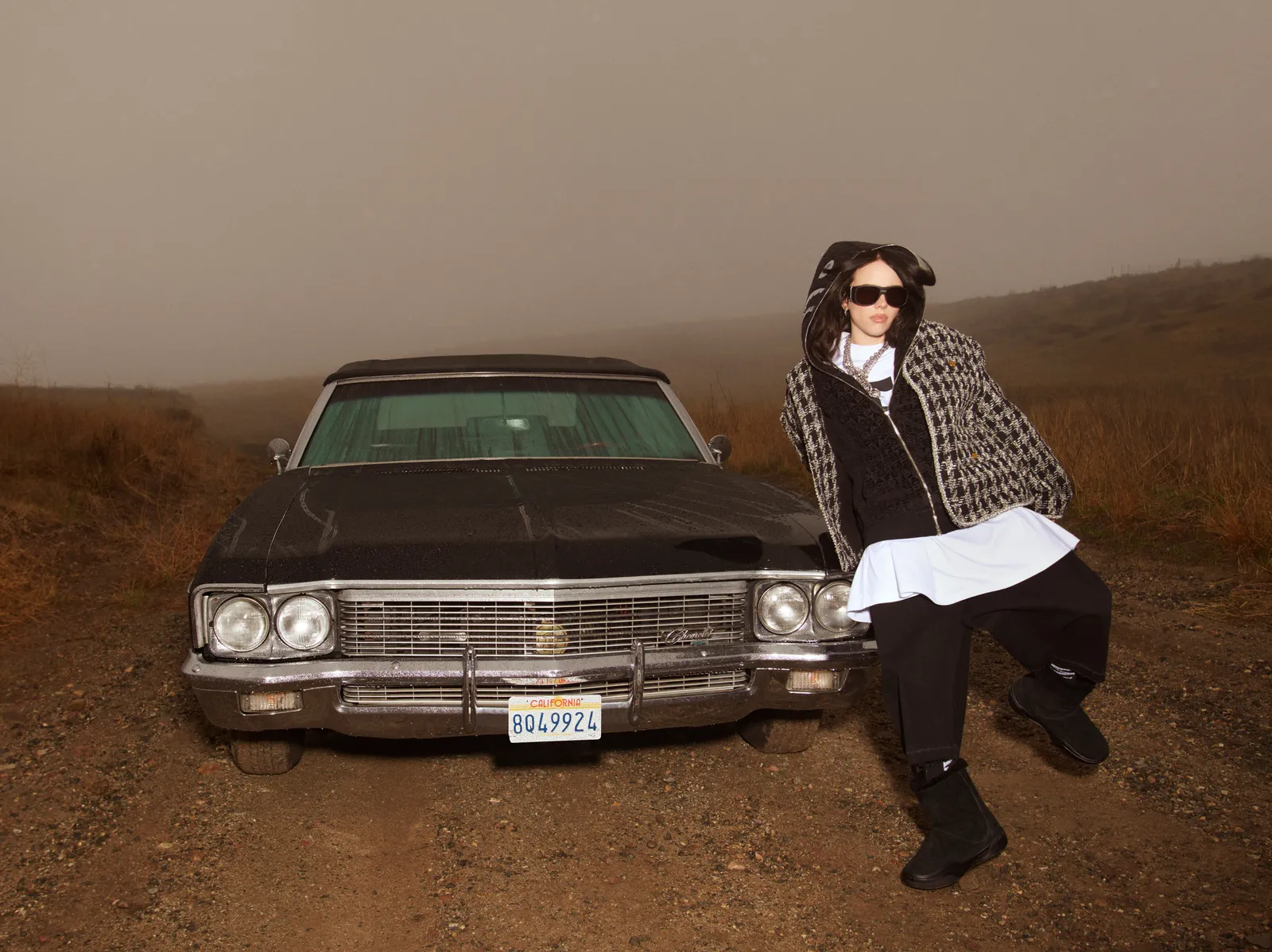 Billie Eilish poses beside a vintage black car in a foggy landscape, wearing layered designer streetwear and sunglasses for British Vogue May 2025.