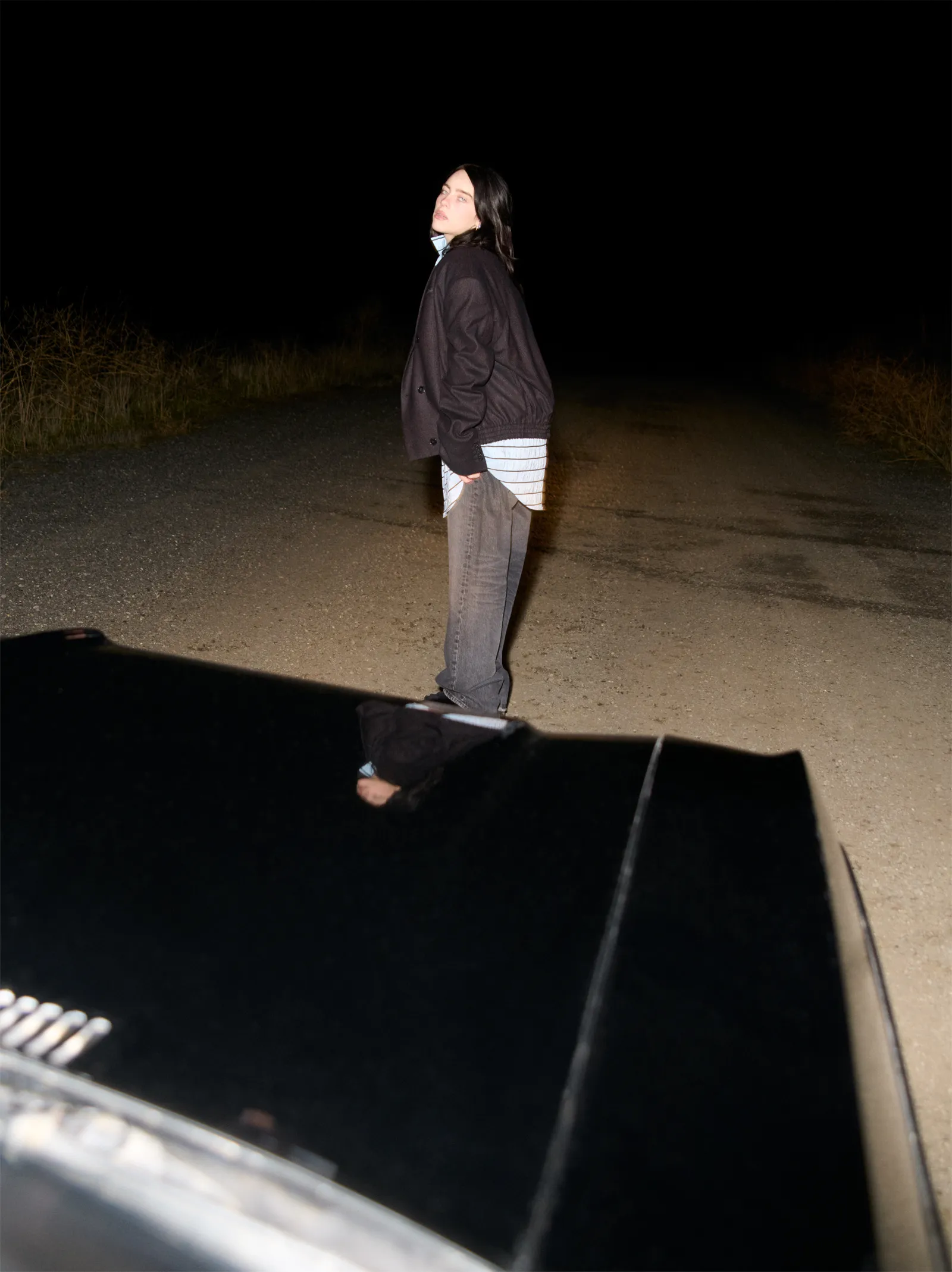 Shot at night, Billie Eilish stands in the middle of a rural road in a striped shirt and dark jacket, illuminated by headlights for British Vogue May 2025.