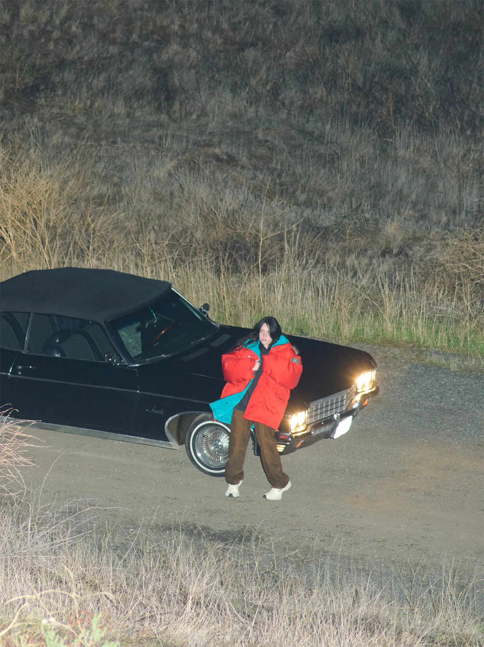 Billie Eilish leans against a black vintage convertible in a bold red and teal puffer jacket during a nighttime desert shoot for British Vogue May 2025.