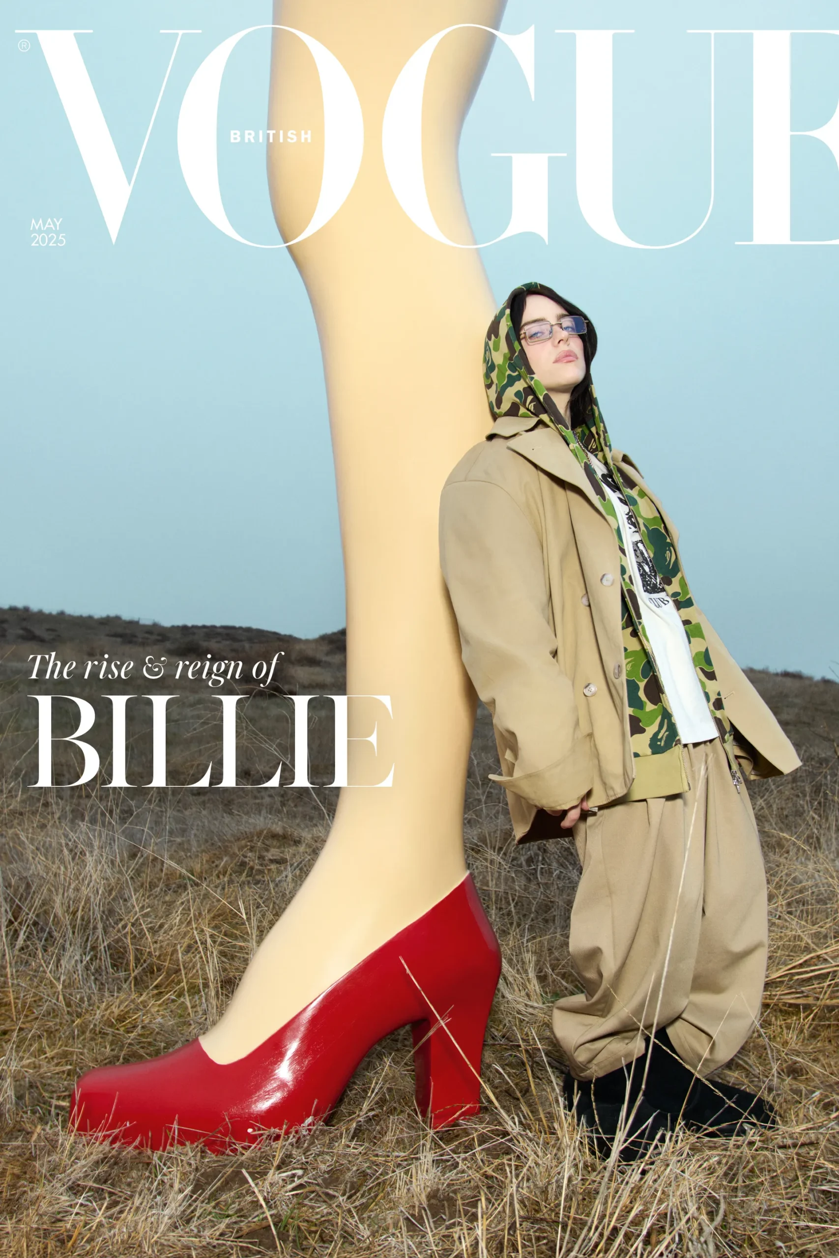 British Vogue May 2025 cover featuring Billie Eilish in oversized tan tailoring and camouflage hoodie, posed beside a surreal giant leg sculpture in the countryside.