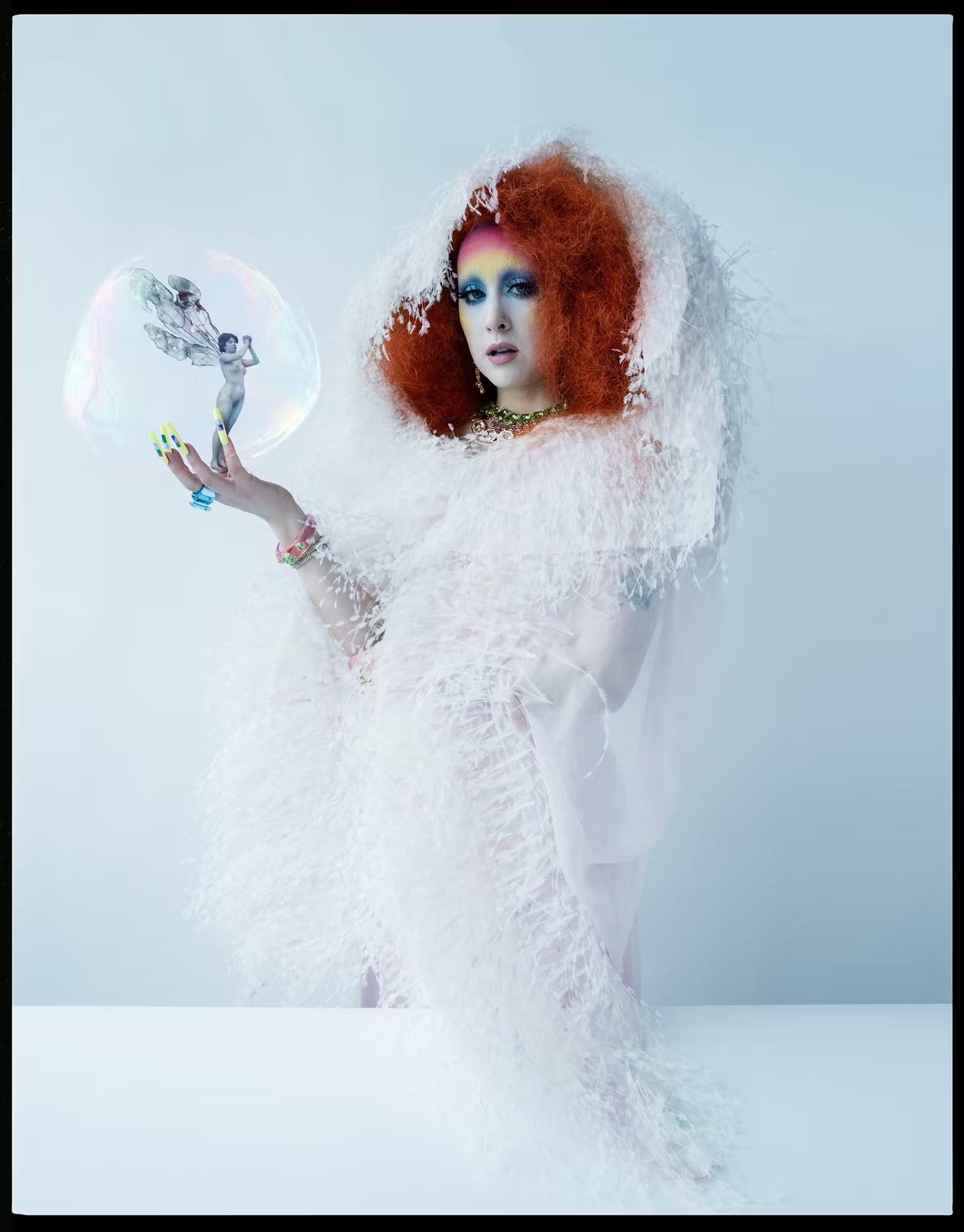 Chappell Roan wears a voluminous white feathered gown with dramatic red hair and rainbow eyeshadow, holding a translucent bubble with a fairy inside – from W Magazine's Pop Issue, May 2025.