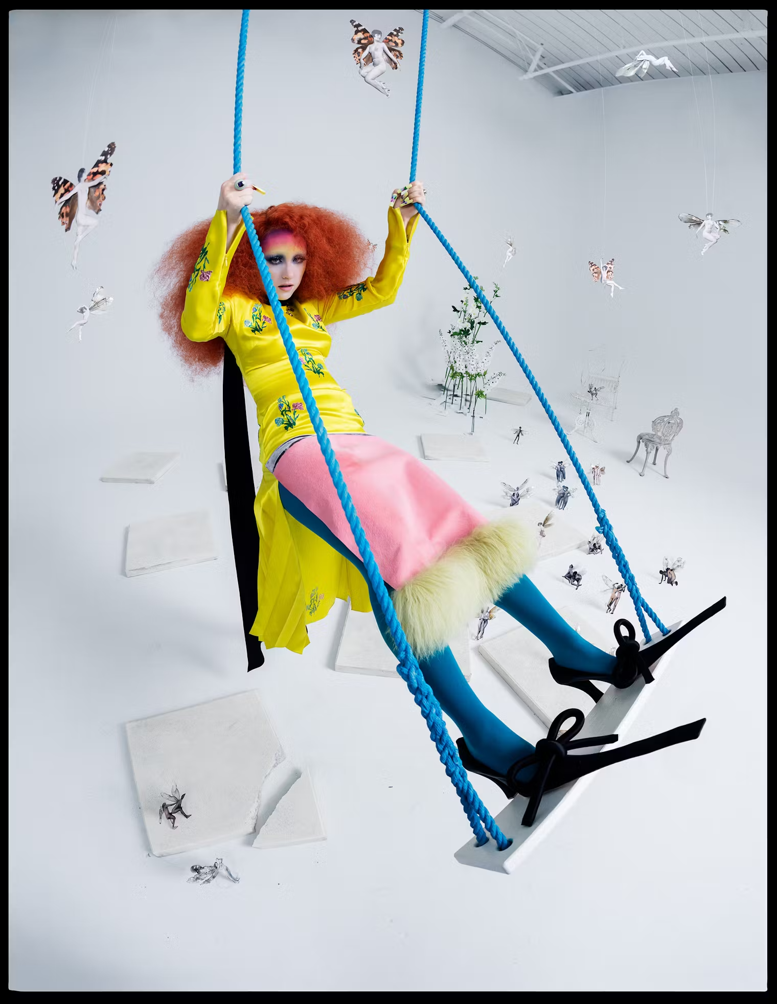 Chappell Roan on a bright blue swing, dressed in a yellow embroidered top, pink fur-trimmed skirt, and teal tights, surrounded by fairy cutouts – W Magazine, May 2025 editorial.