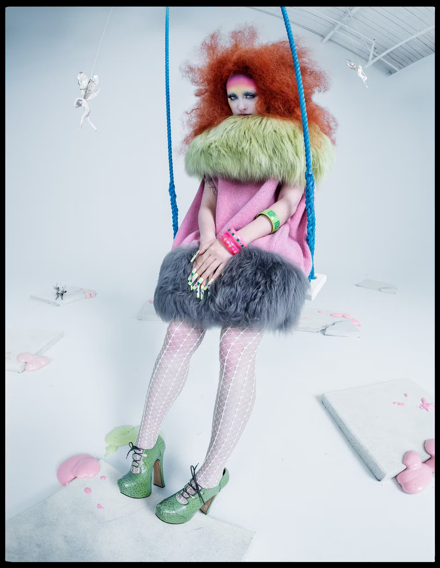 Chappell Roan stands on a swing in a pink and green faux fur dress with fishnet tights and green heels, framed by suspended paper fairies – W Magazine Pop Issue, May 2025.