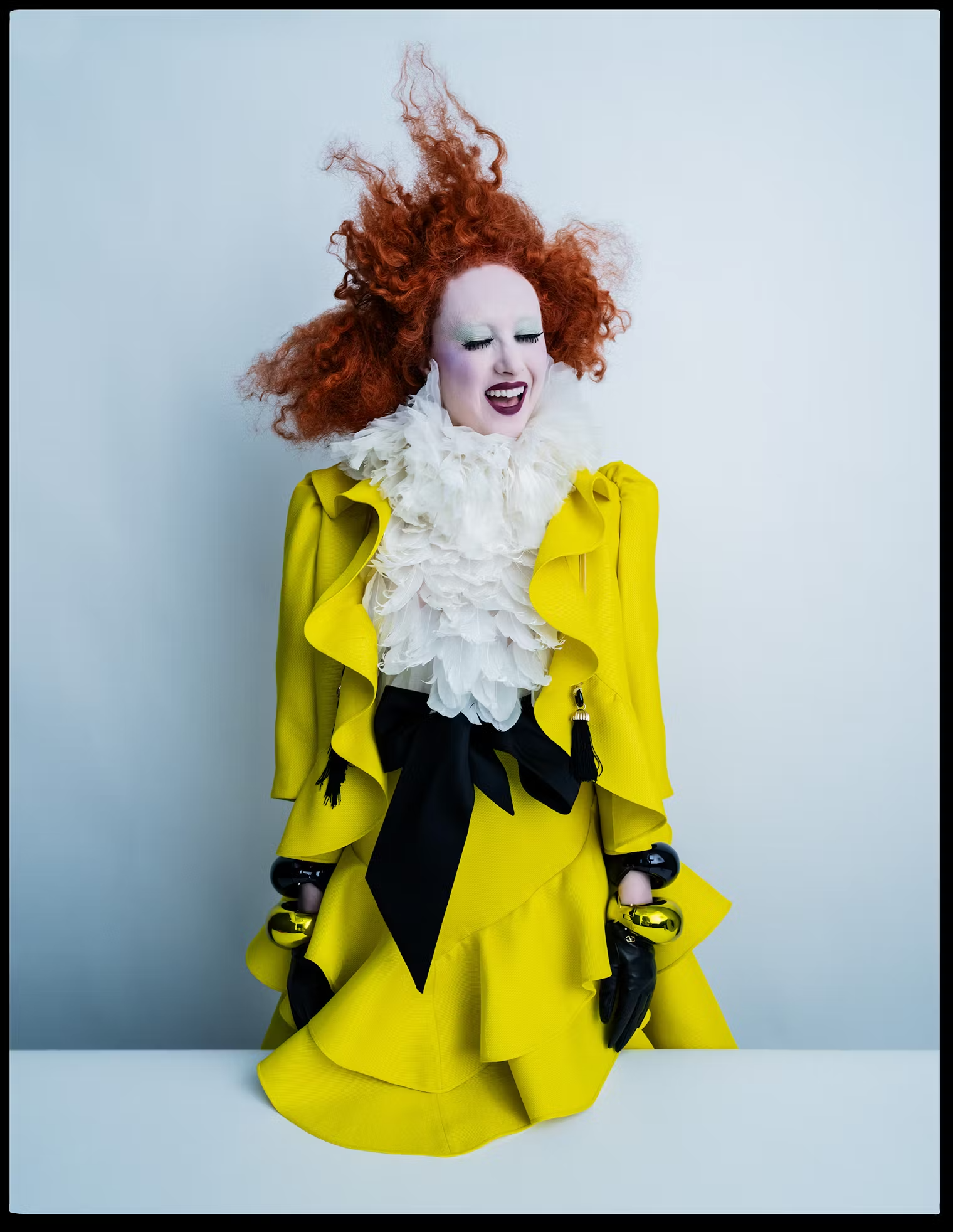 Chappell Roan bursts with laughter in a ruffled yellow outfit with sculpted red curls and dramatic makeup, against a soft blue backdrop – W Magazine Pop Issue May 2025.
