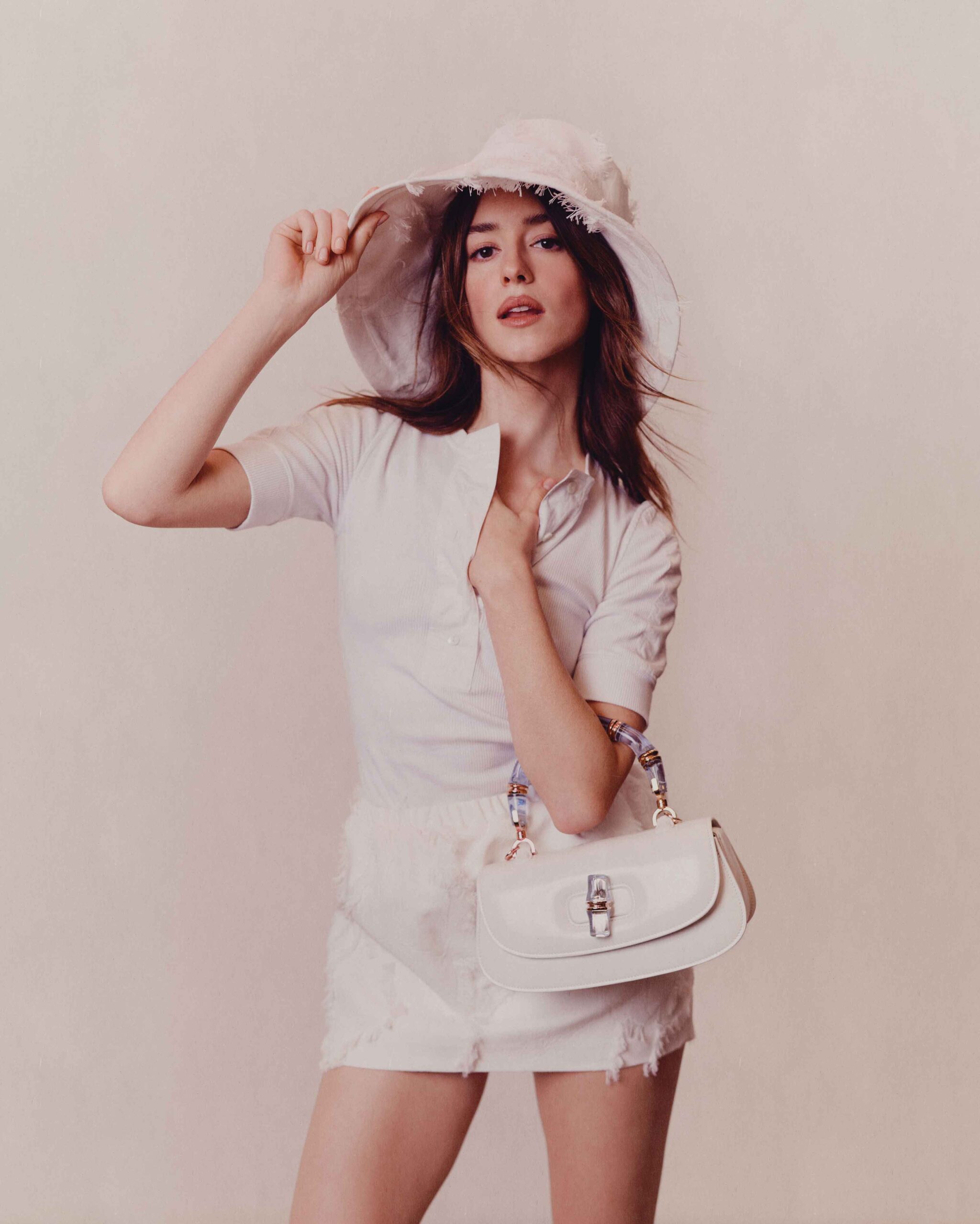 Daisy Edgar-Jones poses in a textured white outfit and matching sunhat, holding a white handbag in a minimalist studio setting.