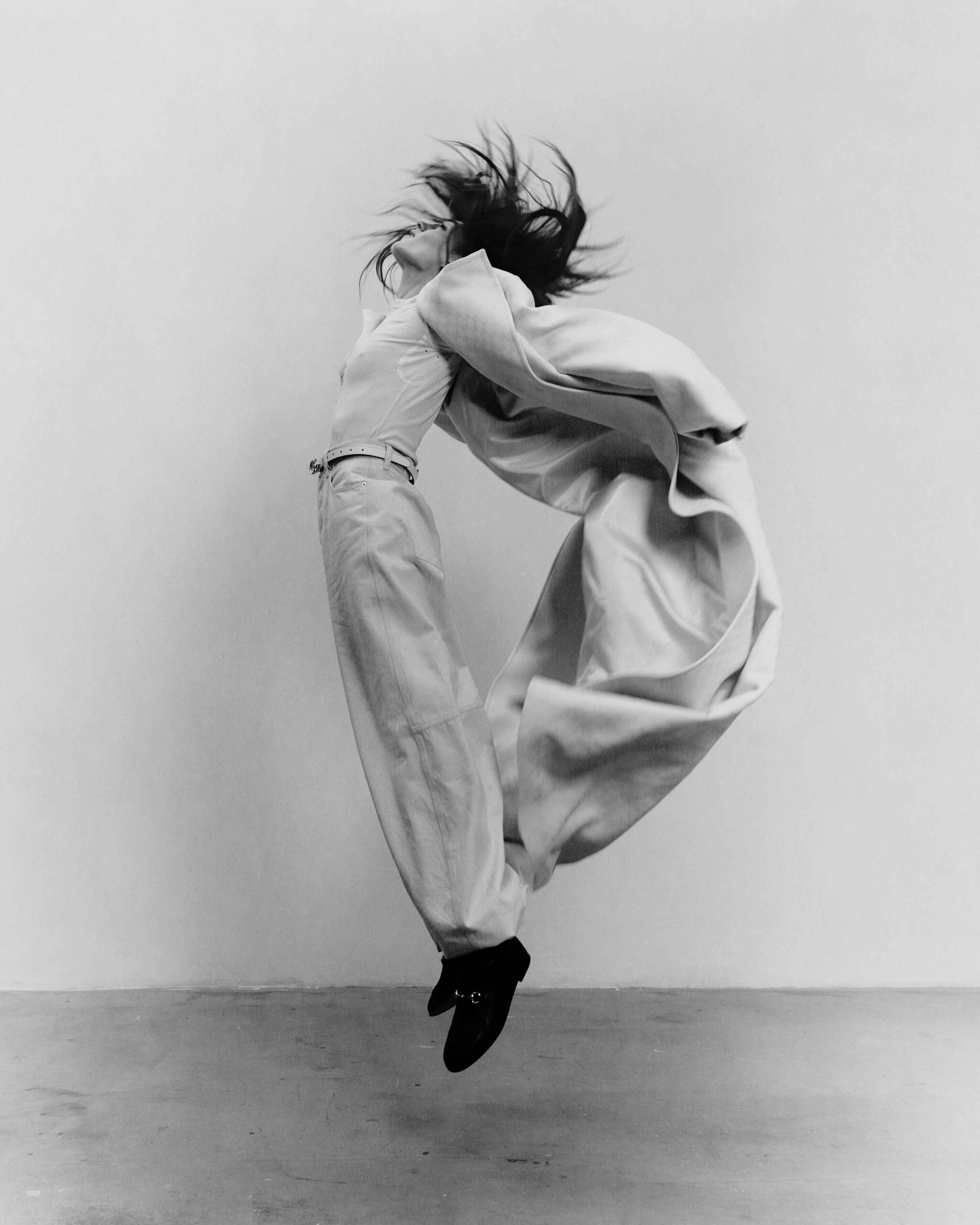 Daisy Edgar-Jones leaps mid-air in wide-leg trousers and a flowing coat, creating a dynamic and dramatic editorial shot.