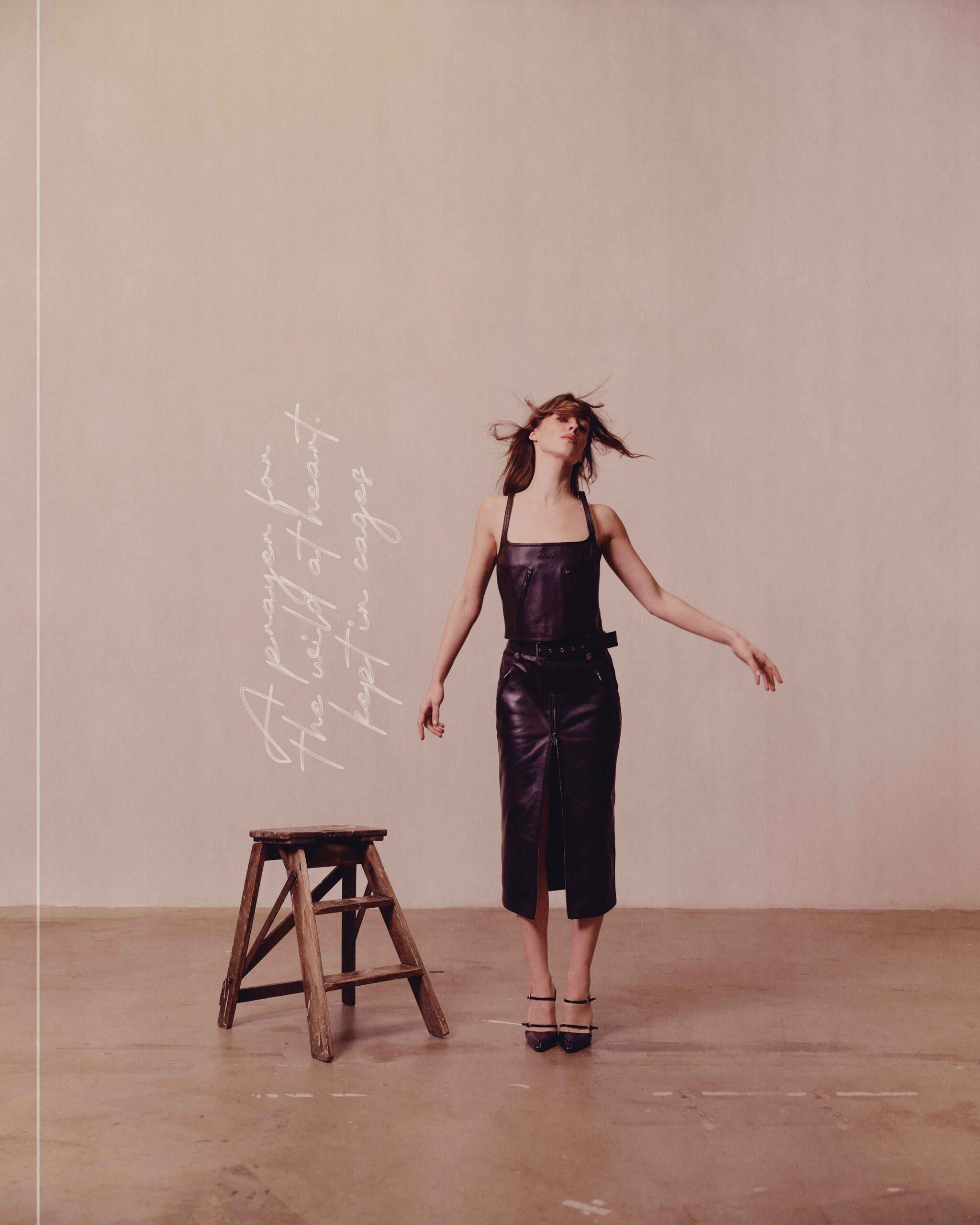 Daisy Edgar-Jones stands beside a wooden stool in a leather dress, with handwritten text on the wall reading “A prayer for the wild at heart kept in cages.”