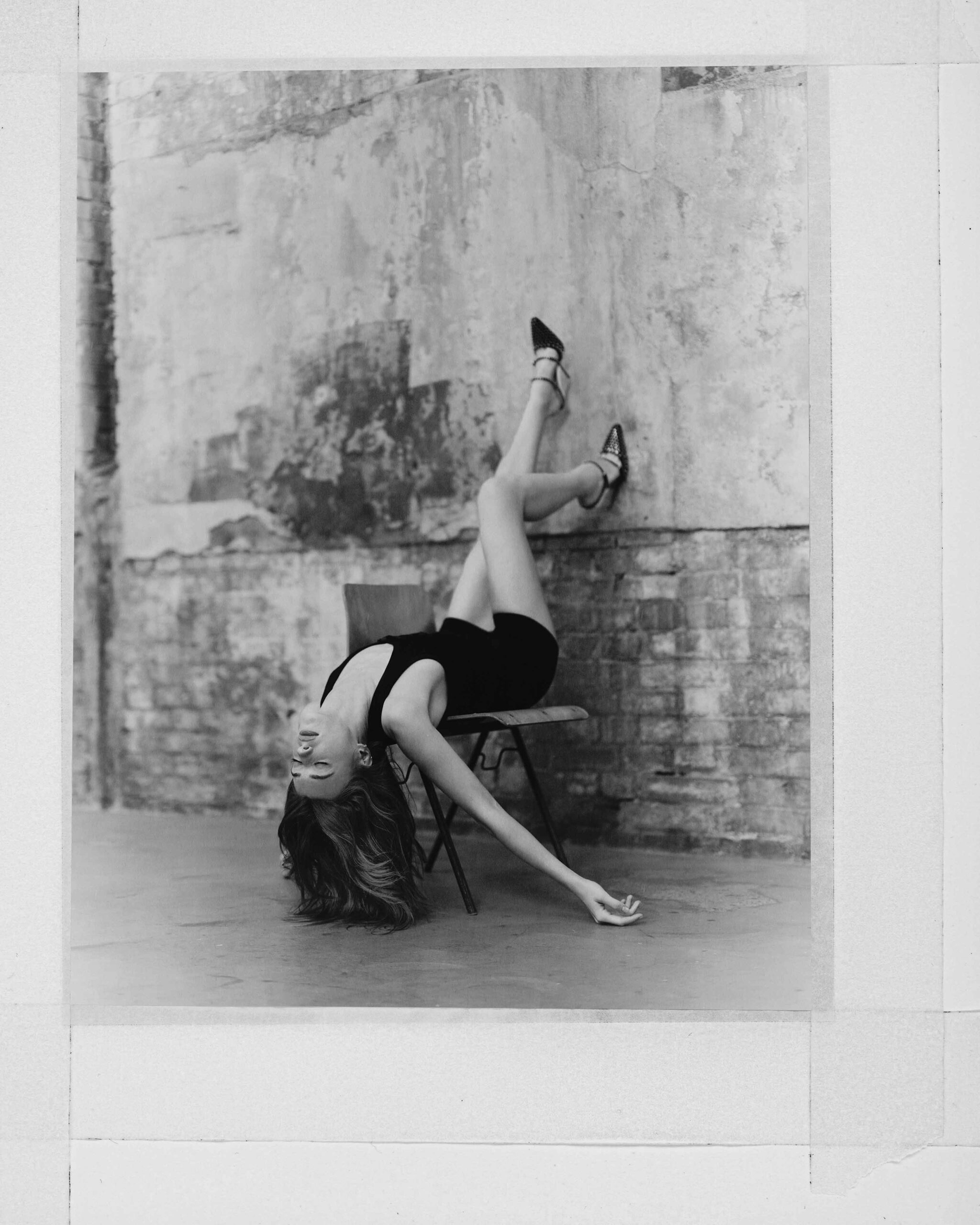 Daisy Edgar-Jones reclines dramatically on a chair against a textured brick wall, wearing a black bodysuit and heels in a black-and-white shot.