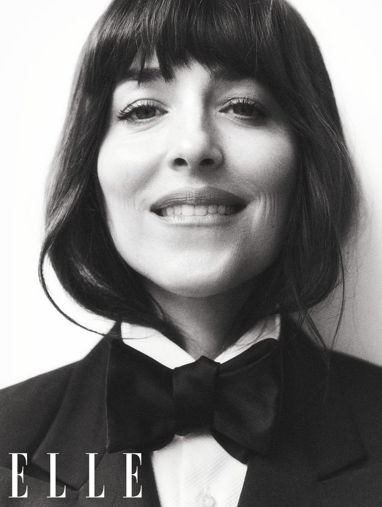 Close-up portrait of Dakota Johnson in a tuxedo and bow tie, smiling softly in a black and white cover for ELLE UK June 2025.