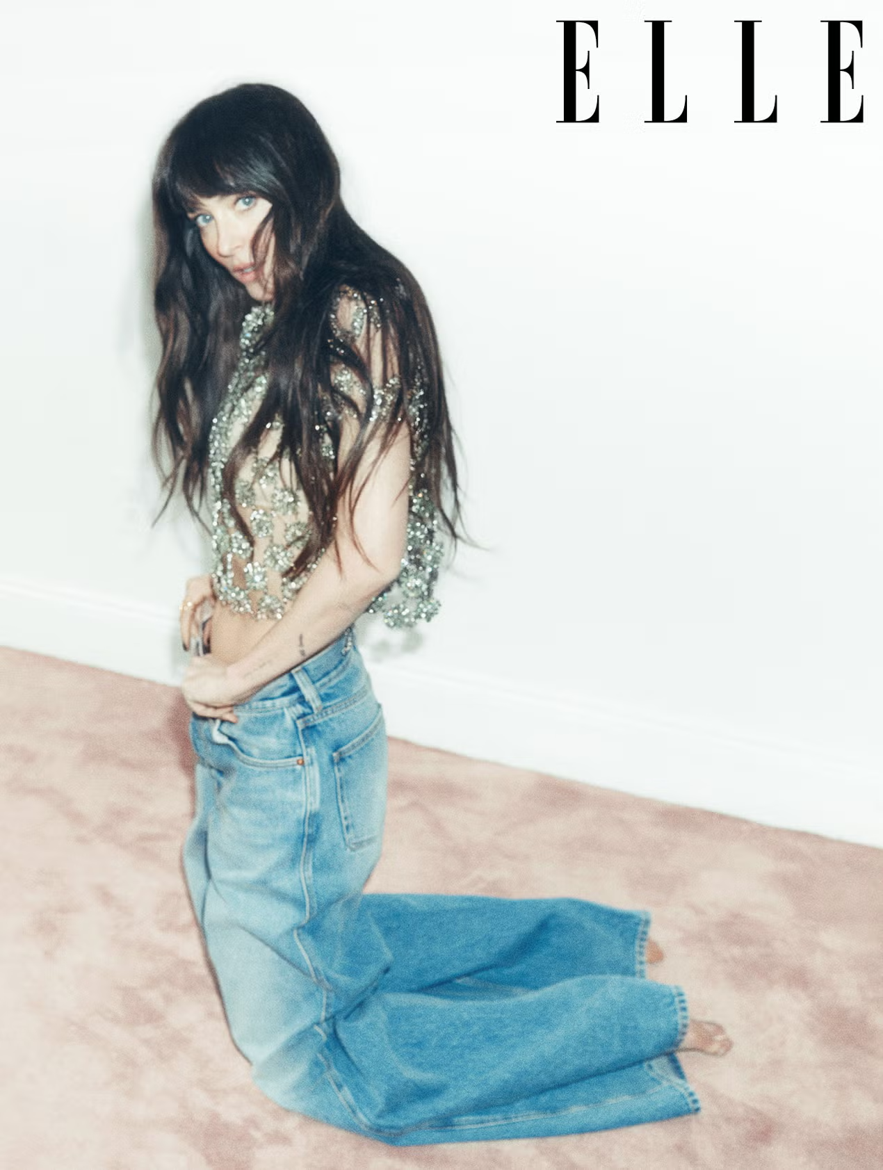 Dakota Johnson kneeling on a pink carpet in oversized jeans and an embellished sheer top, styled for ELLE UK.