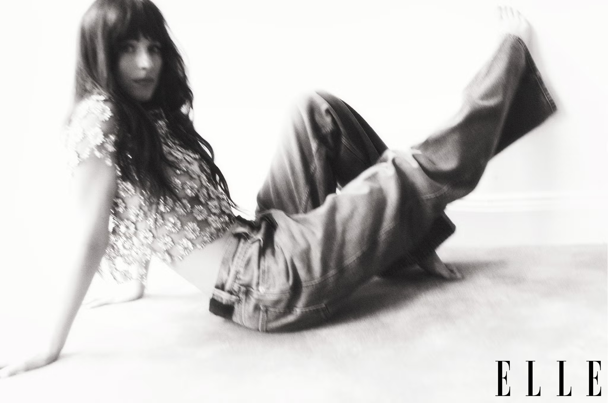 A blurred black-and-white image of Dakota Johnson reclining in floral top and denim jeans for ELLE UK June 2025.