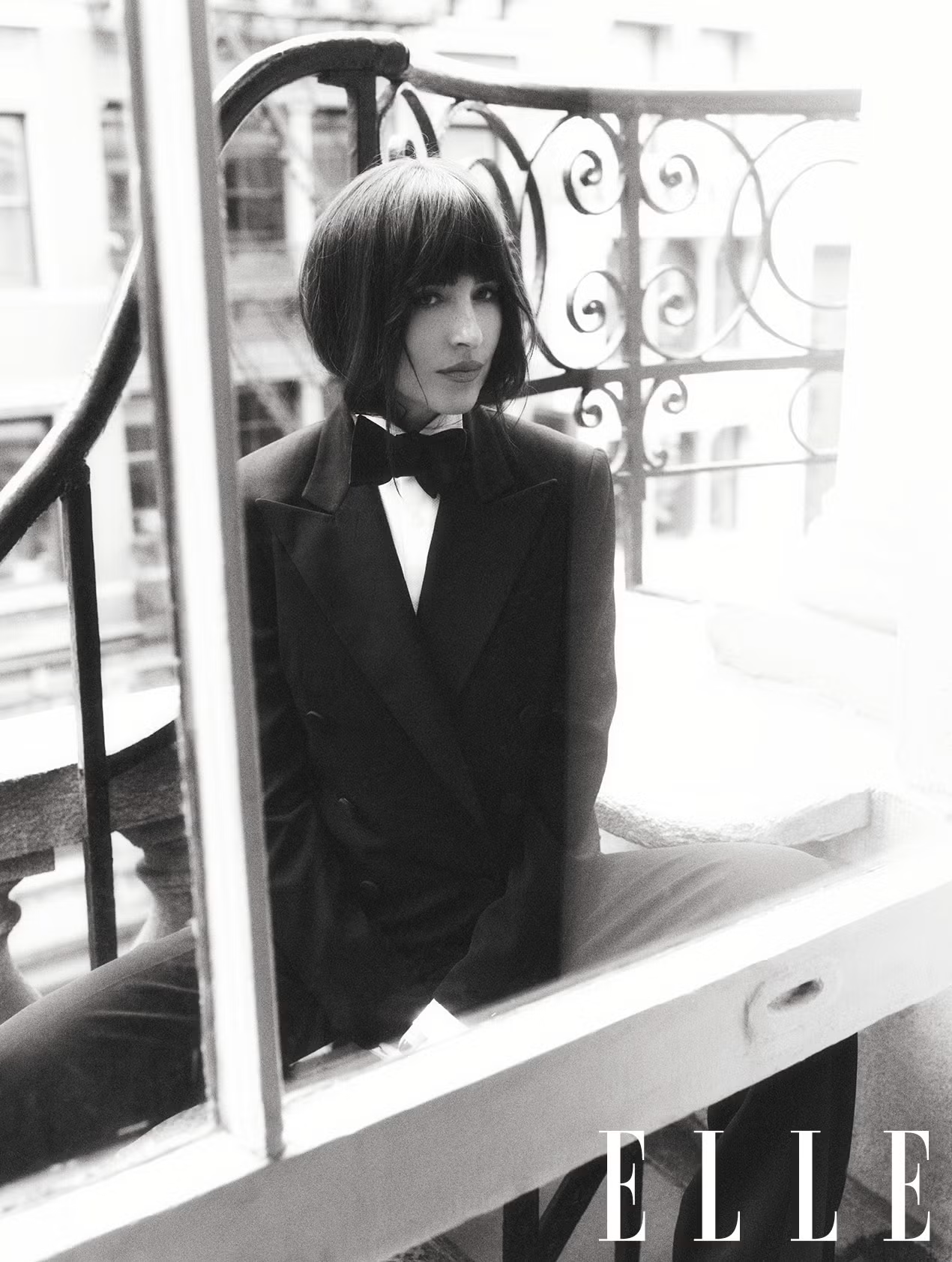 Side profile of Dakota Johnson sitting on a balcony in a tailored suit and bow tie, styled in classic black and white.