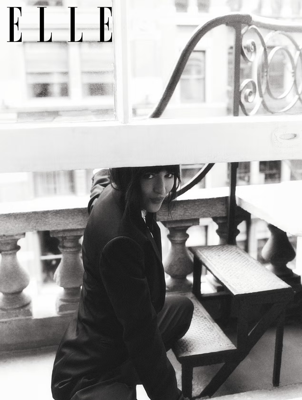 Dakota Johnson kneeling on a balcony stairway in a formal suit, looking up with an intense gaze, featured in ELLE UK.