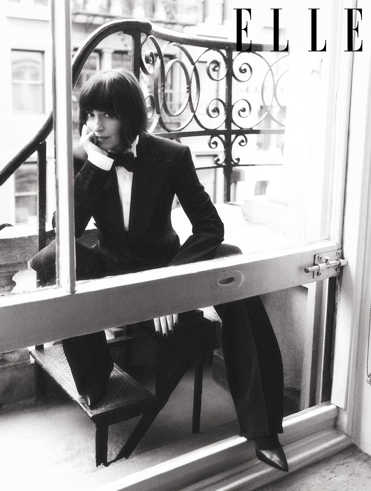 Dakota Johnson seated on a balcony in a black tuxedo and stilettos, gazing through a window for ELLE UK June 2025.
