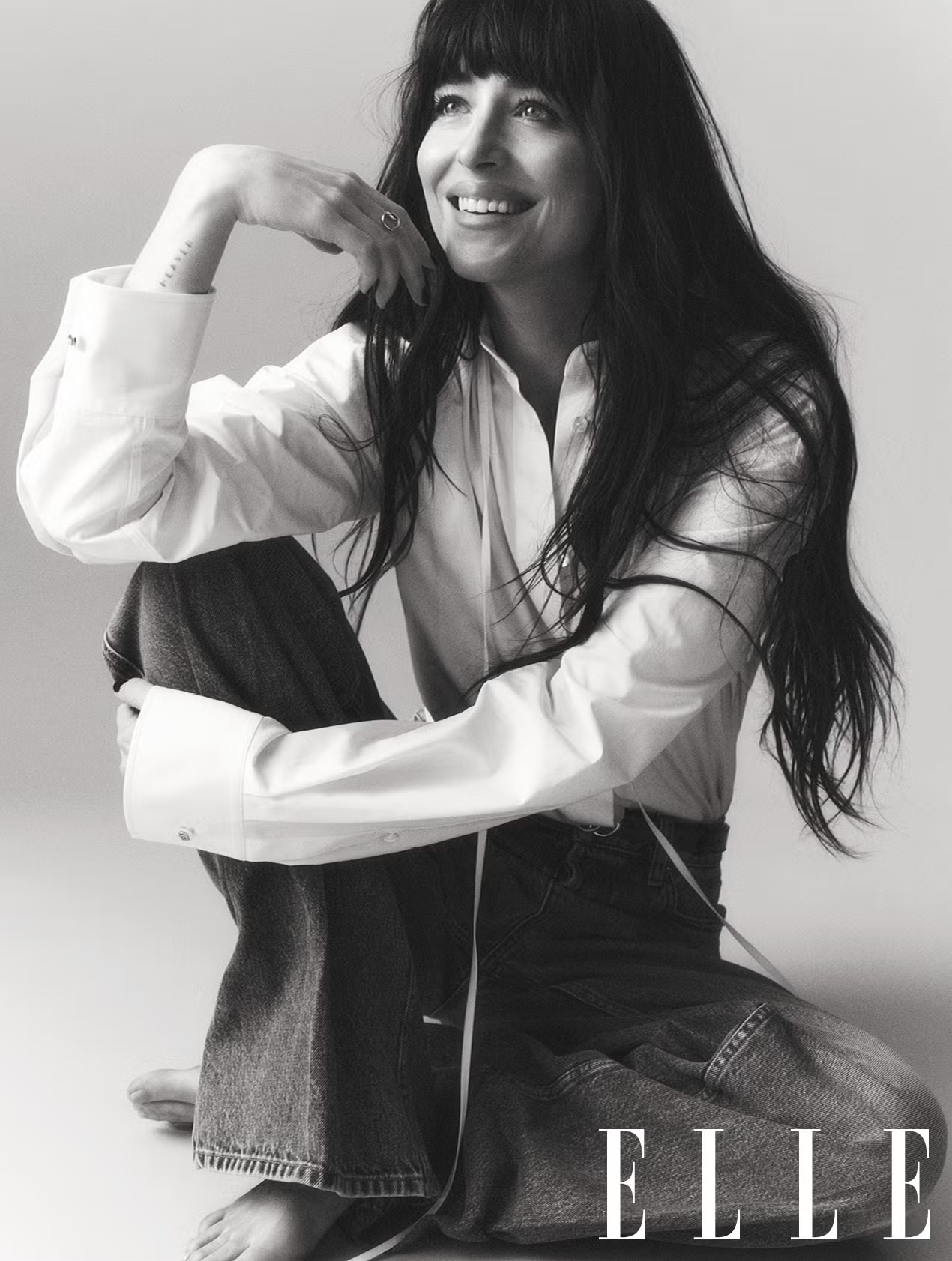 Dakota Johnson in a relaxed pose on the floor in jeans and a white blouse, smiling naturally for ELLE UK.