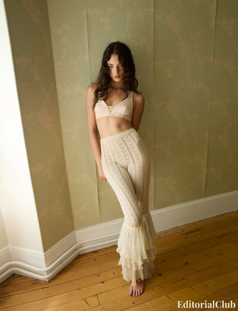 Deva Cassel poses indoors in a soft pink bralette and vintage-inspired flared knit pants with ruffle hems.