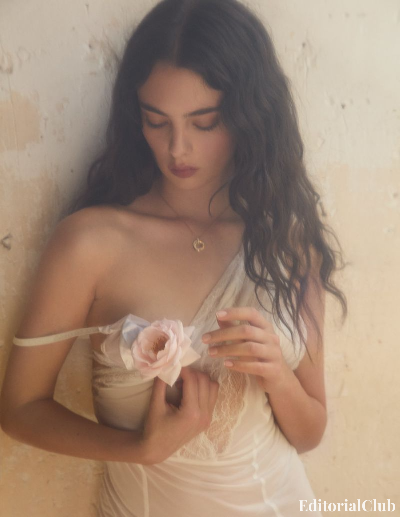 Deva Cassel gazes down at a soft pink flower while wearing a sheer lace gown in a sun-drenched room.