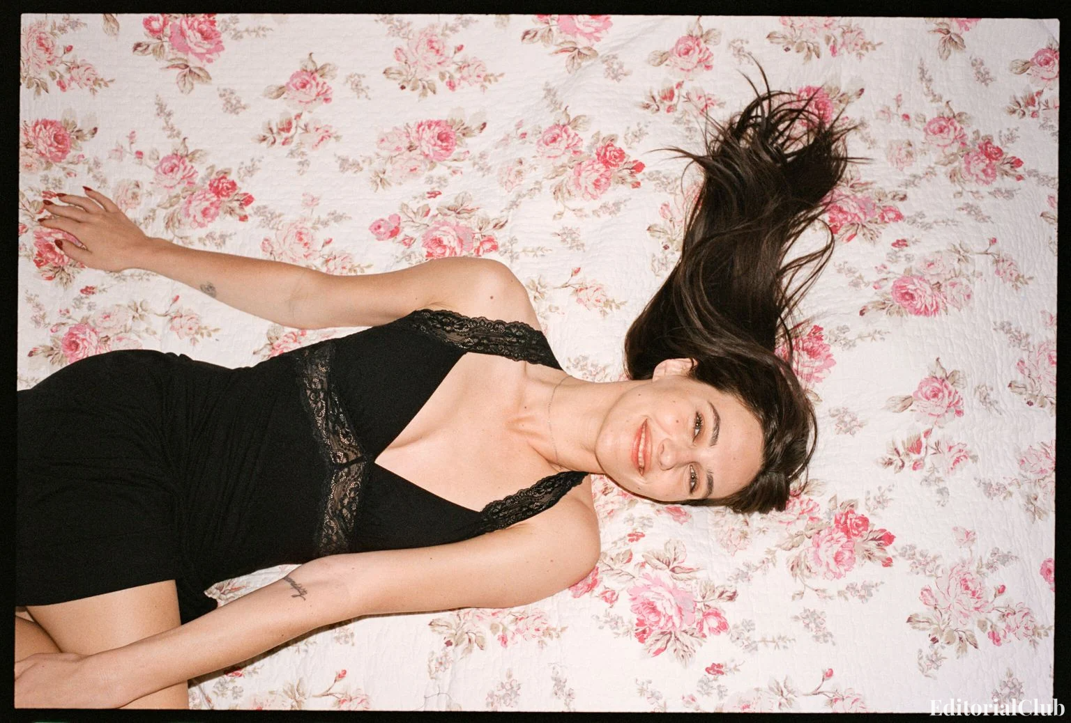 Devon Lee Carlson smiling while lying on a floral bedspread in a black lace dress — Reformation 2025 editorial.