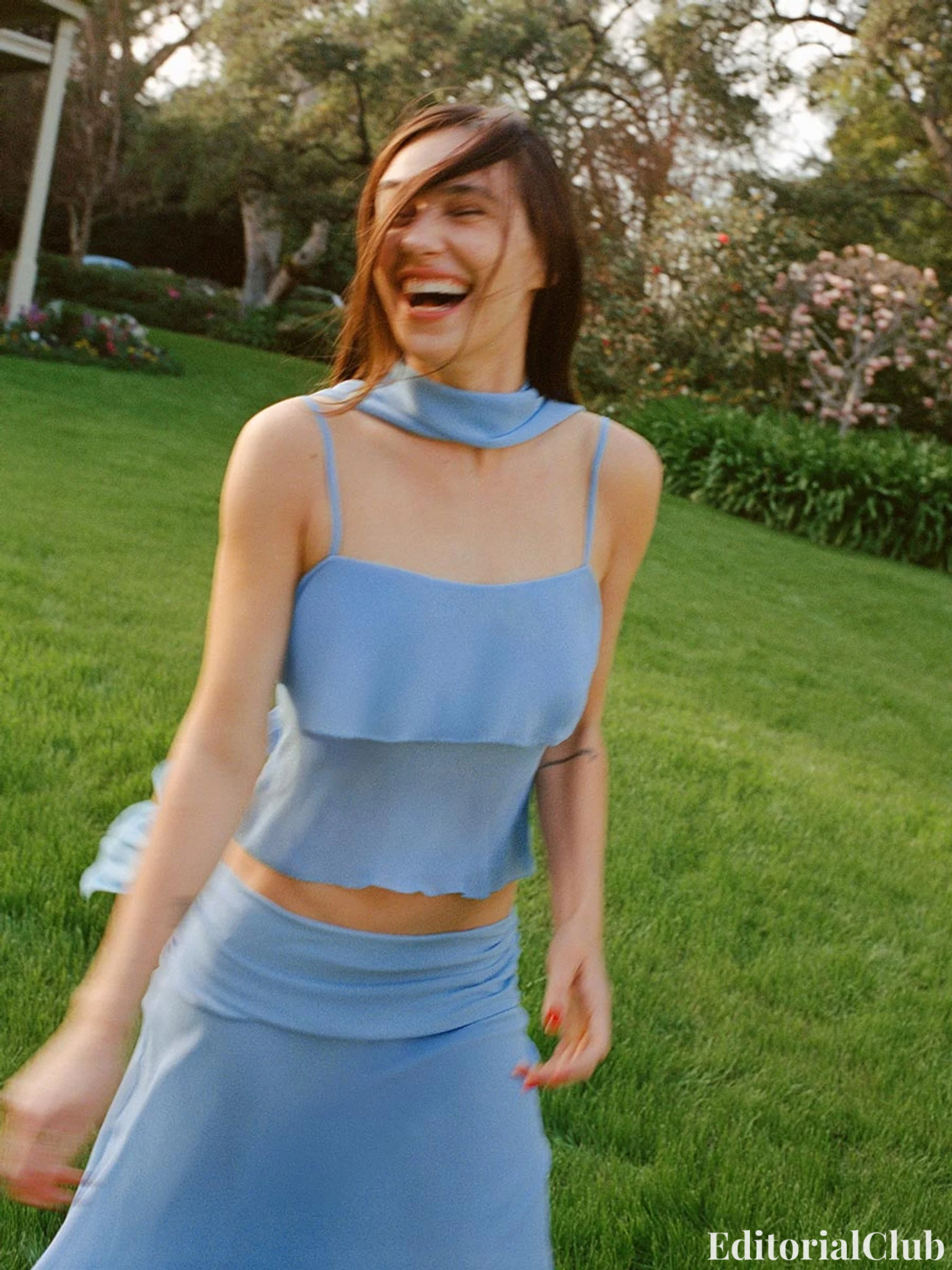 Devon Lee Carlson laughs mid-spin in a flowing blue outfit on a garden lawn — Reformation Spring 2025.