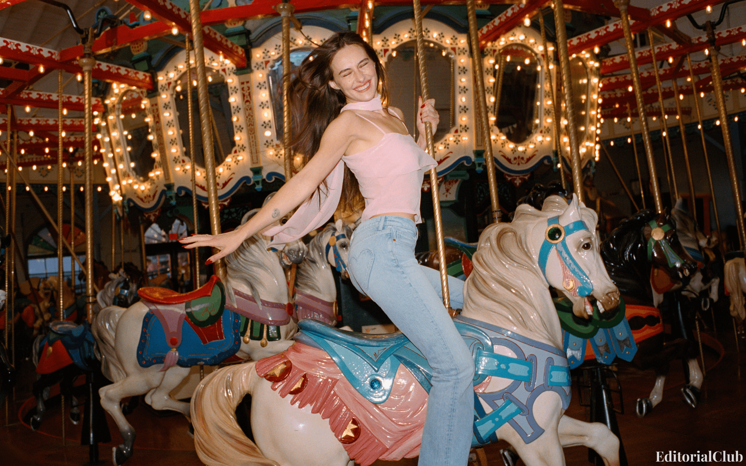 Devon Lee Carlson rides a carousel horse in jeans and a pink top, radiating fun and retro vibes — Reformation 2025.