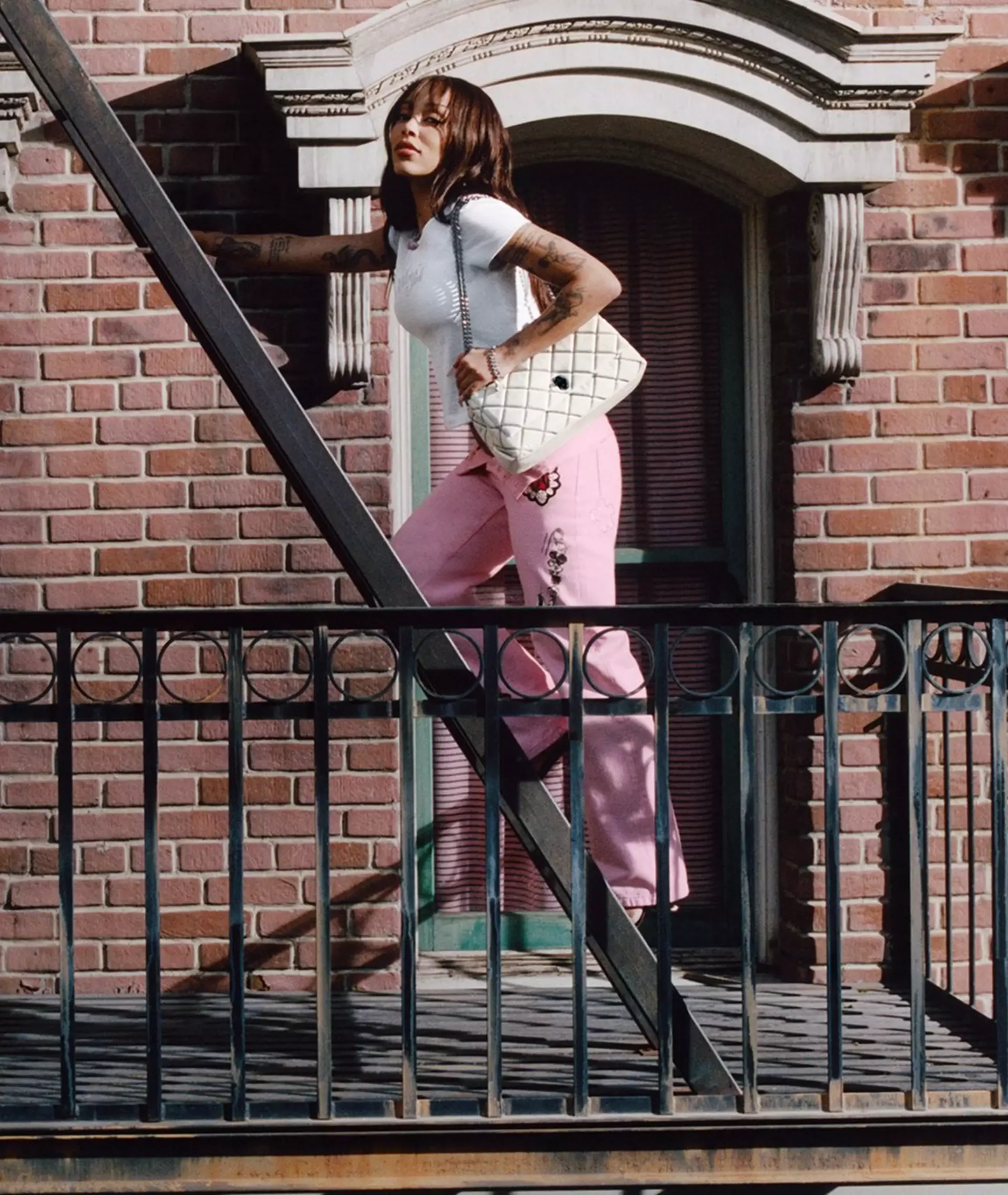 Doja Cat walking up a fire escape in pink pants and a white fitted t-shirt, carrying a quilted white Marc Jacobs shoulder bag