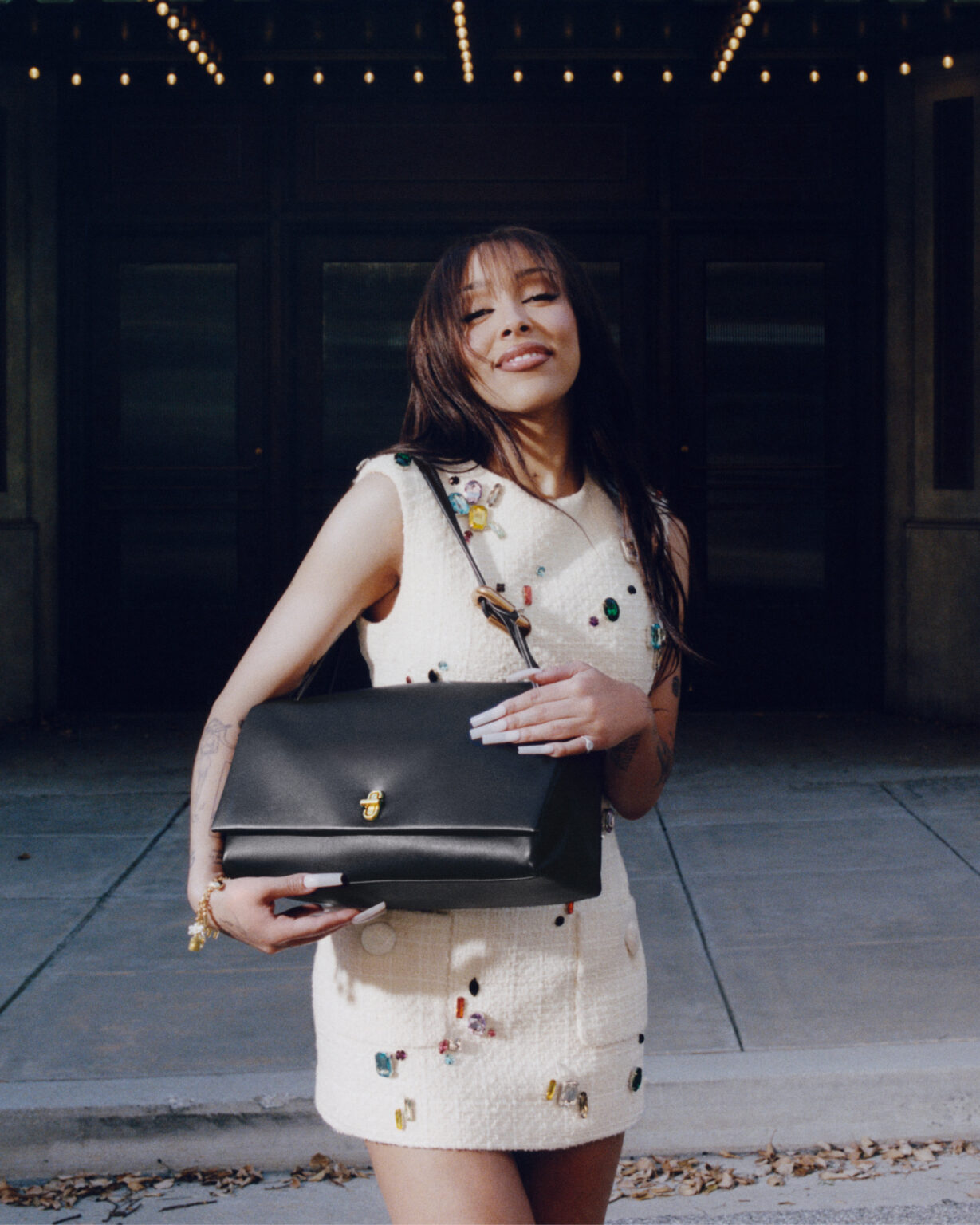 Doja Cat posing with a black leather Marc Jacobs handbag while wearing a white embellished mini dress in a city setting