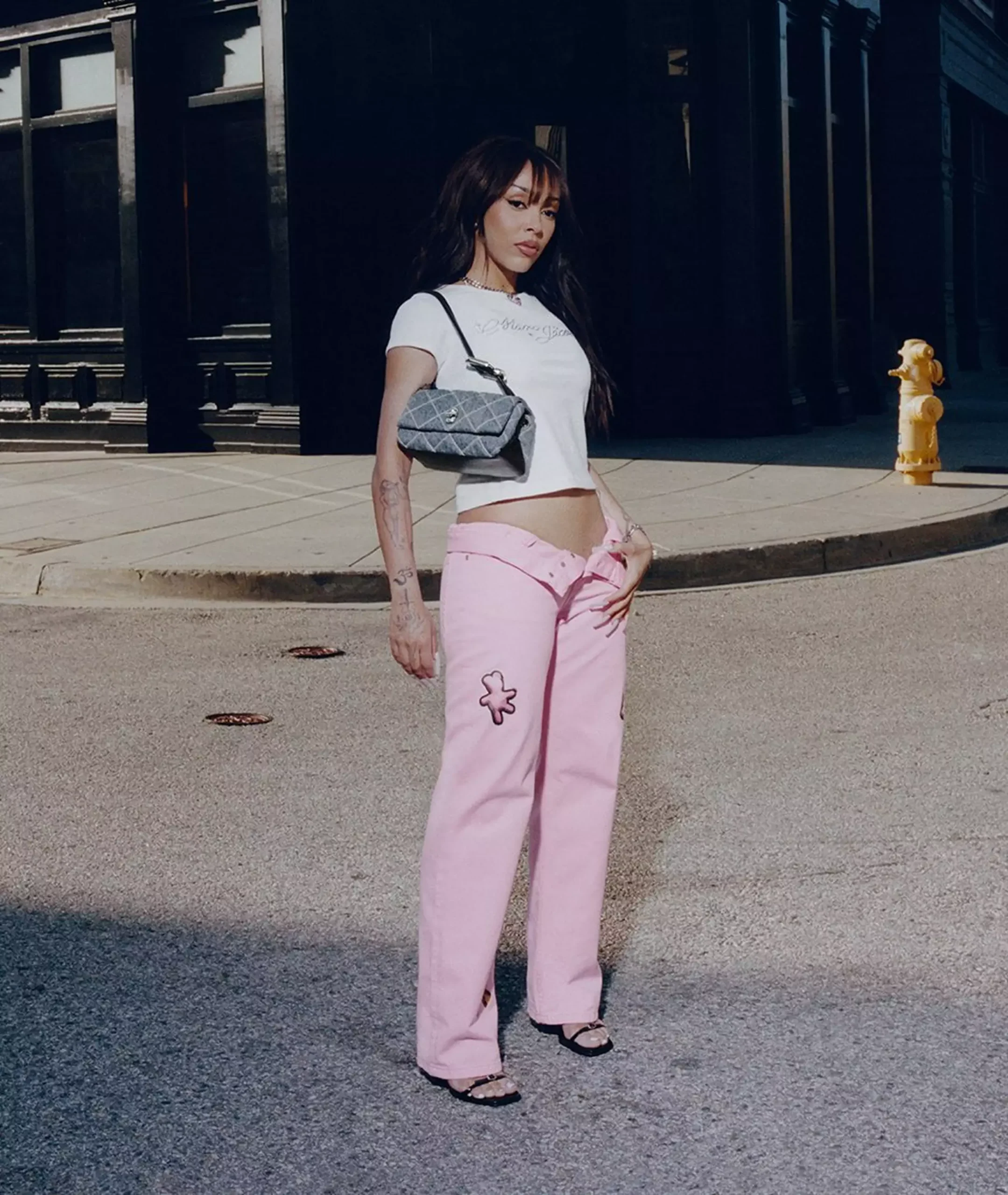 Doja Cat standing in the street wearing low-rise pink jeans and a cropped white tee, paired with a quilted grey shoulder bag