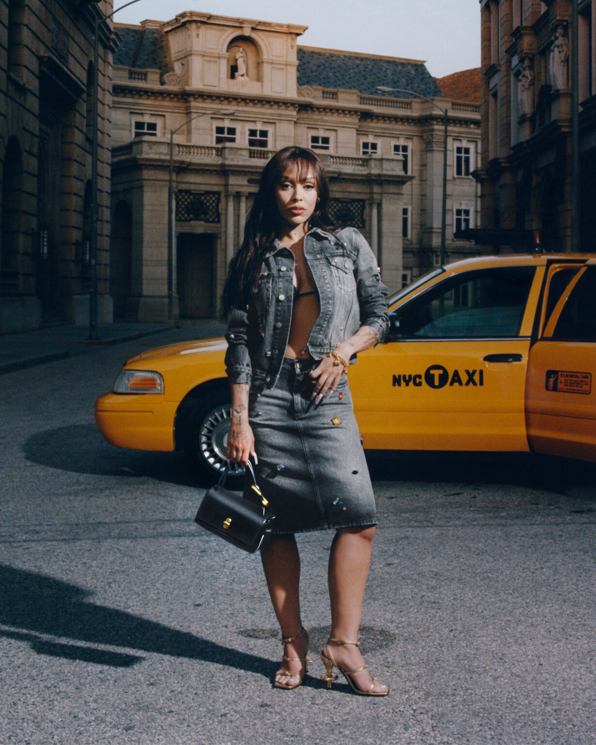 Doja Cat in a grey denim co-ord set with a black purse, posing confidently in front of a classic NYC yellow taxi
