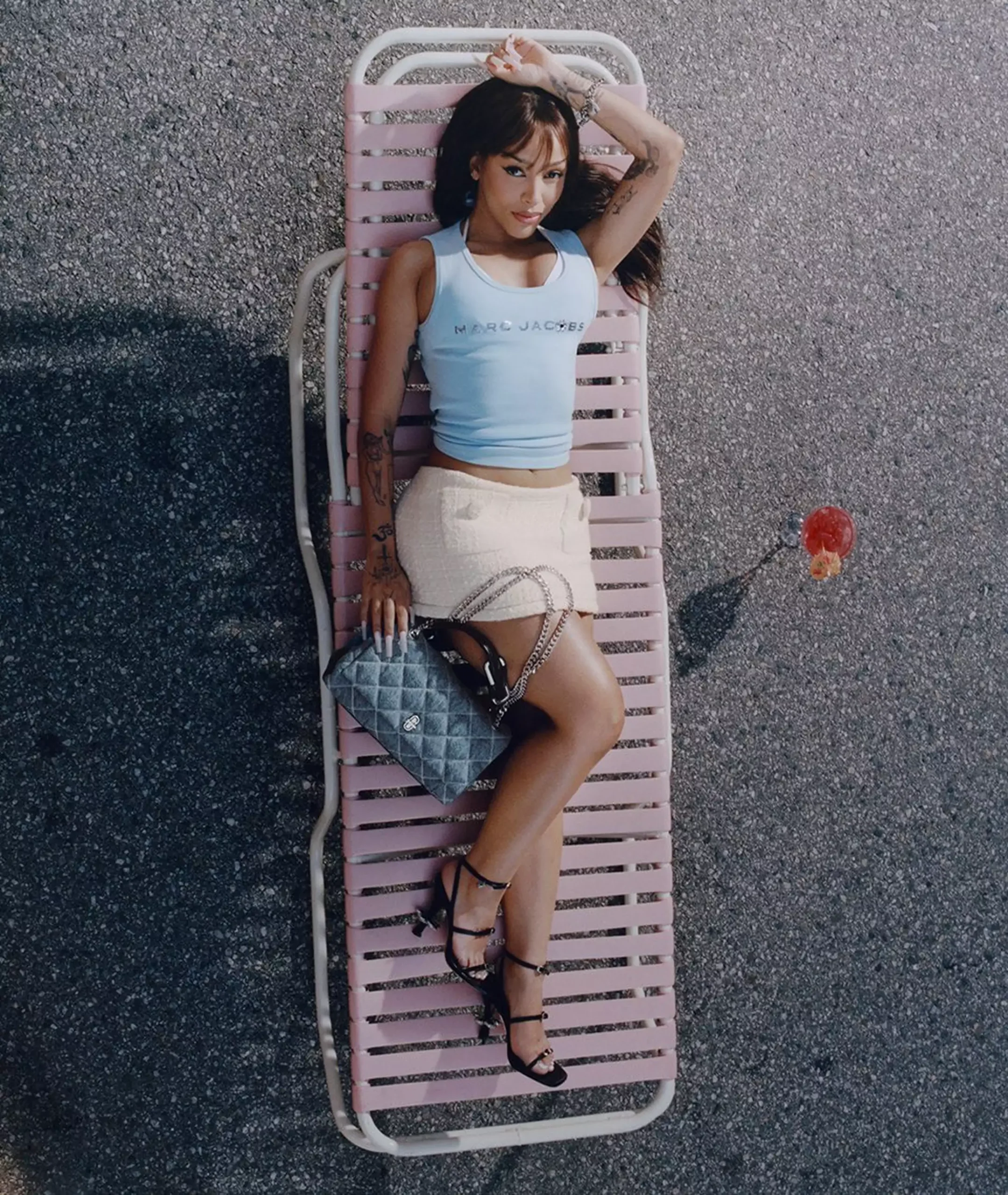 Doja Cat lounging on a pink recliner in a light blue tank top and white mini skirt, holding a large grey Marc Jacobs quilted bag