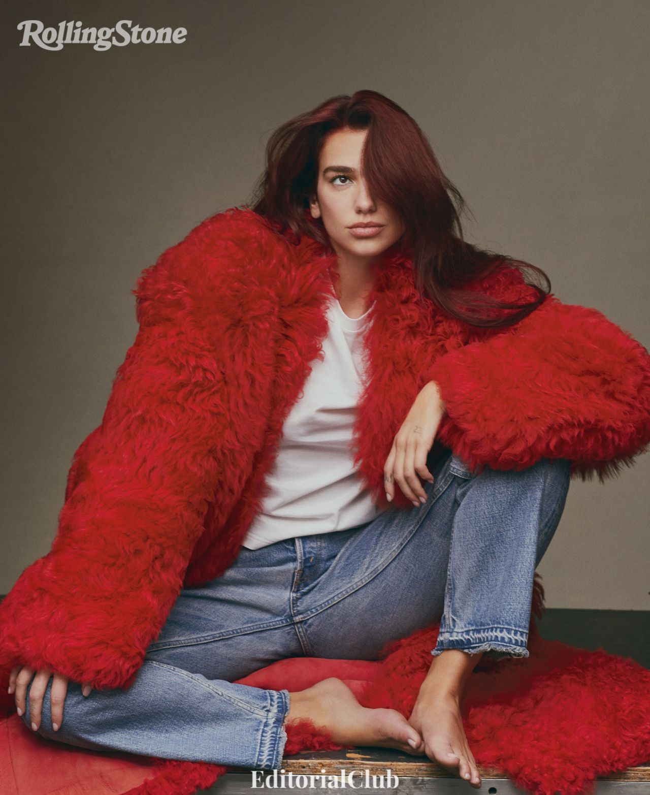 Dua Lipa in red faux fur coat and jeans, barefoot on set for Rolling Stone January 2024 editorial.