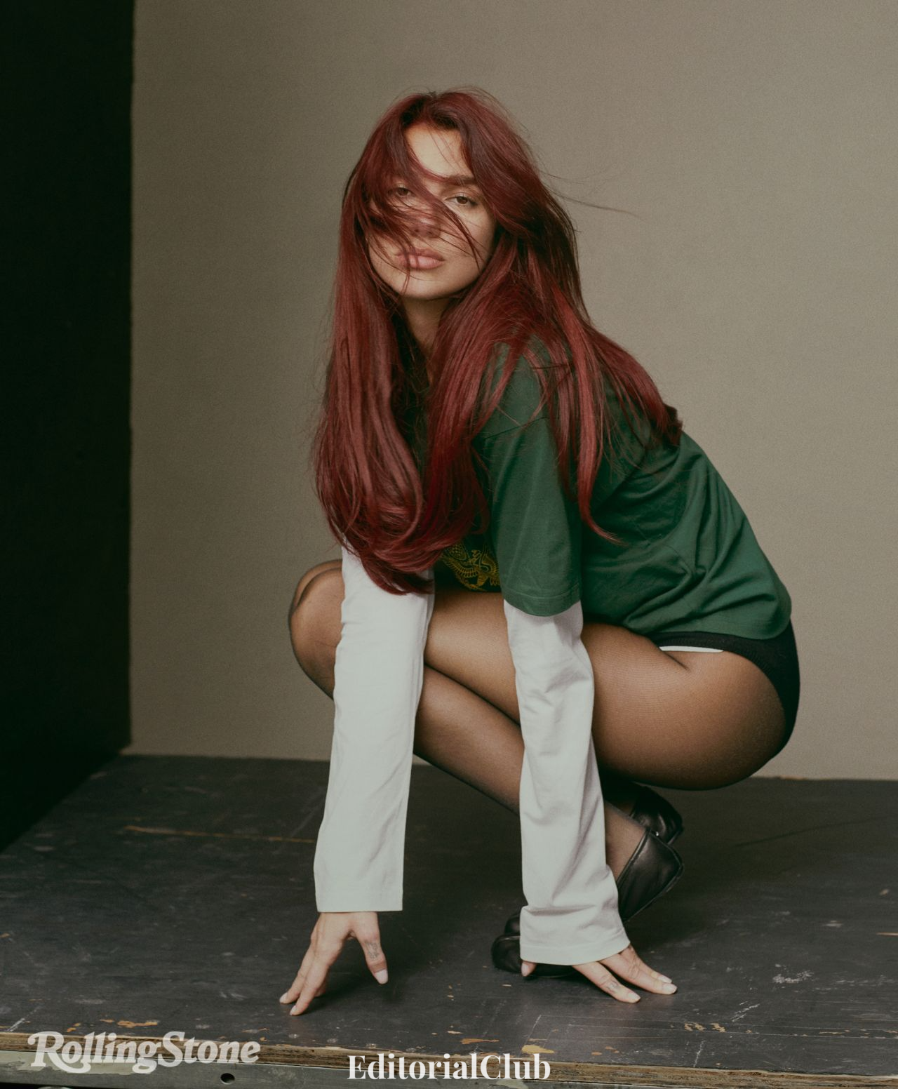 Dua Lipa crouching in green and white layered shirt with tousled red hair during Rolling Stone photoshoot.