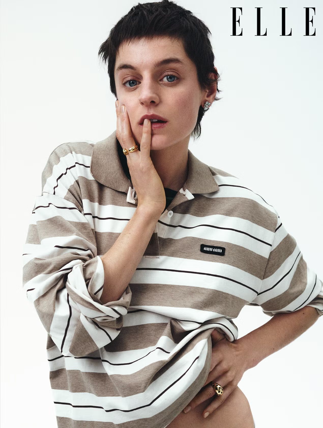 Emma Corrin poses in striped Miu Miu rugby shirt for ELLE UK April 2025 cover shoot, styled with gold rings and minimal makeup