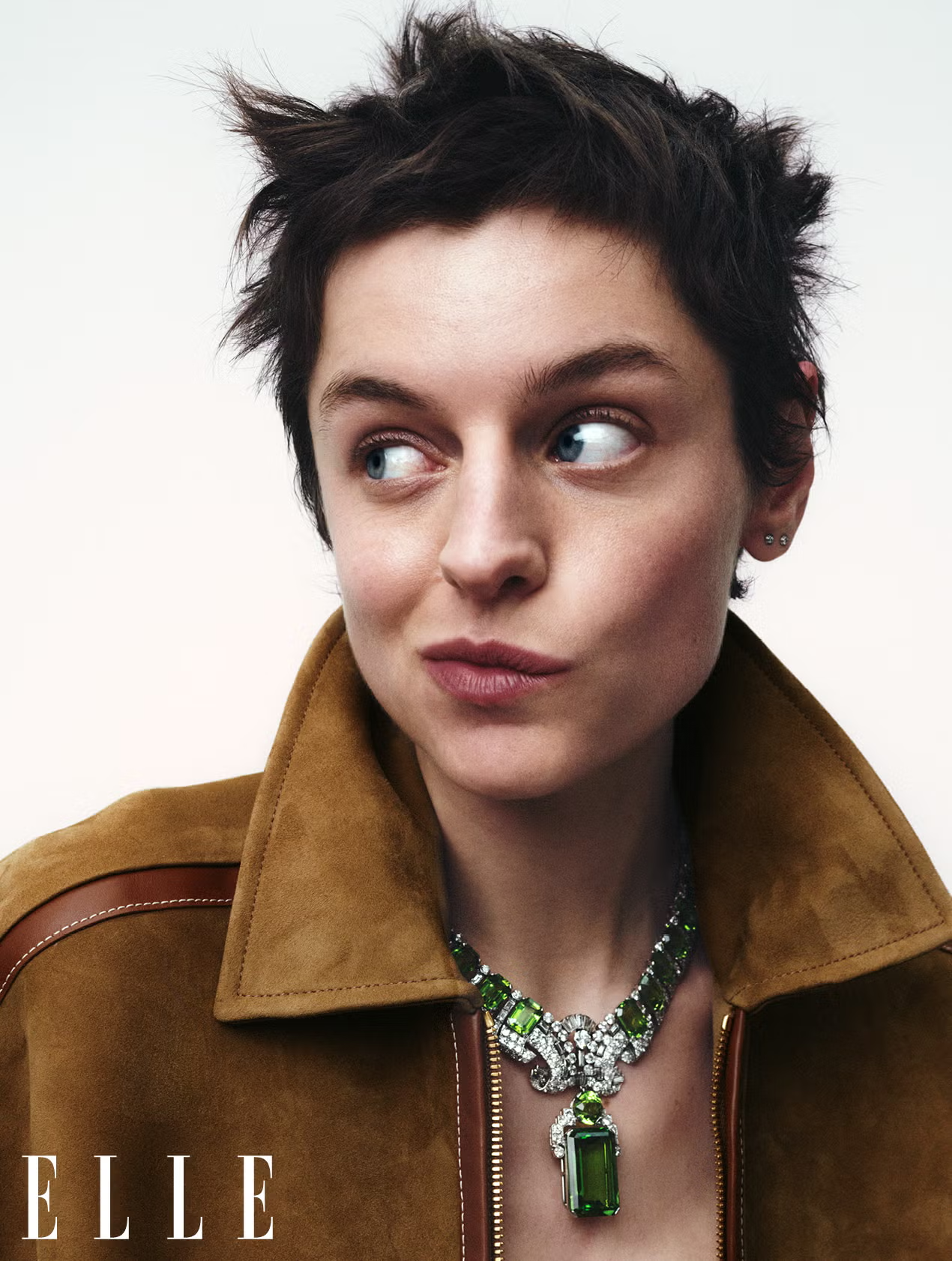Close-up portrait of Emma Corrin in brown suede jacket with bold green gemstone necklace for ELLE UK editorial
