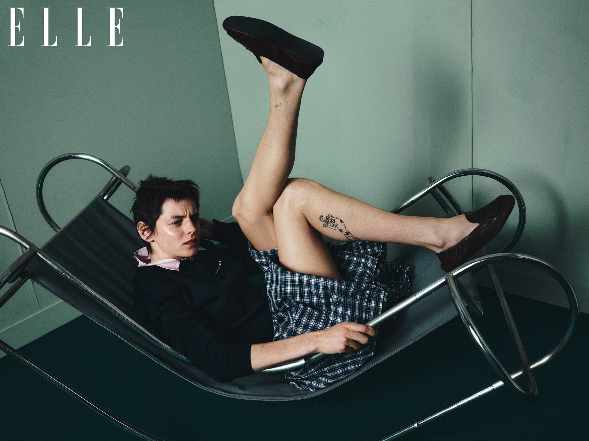 Editorial image of Emma Corrin reclining in a modern metal sling chair, wearing plaid skirt and loafers for ELLE UK
