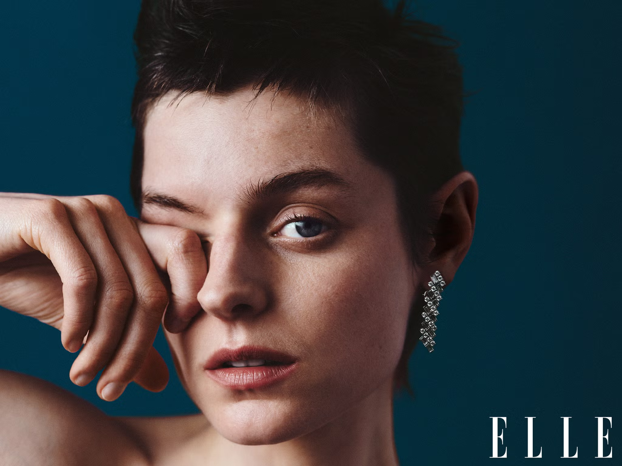 Close-up of Emma Corrin wiping her eye, wearing crystal earrings, for a raw beauty portrait in ELLE UK April 2025
