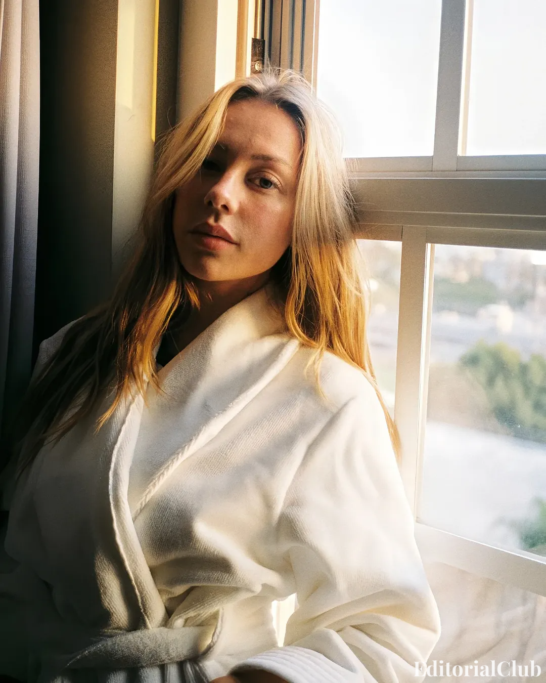 Ester Expósito in a white bathrobe standing by a window with soft morning light on her face during the Not a Doll Desigual campaign