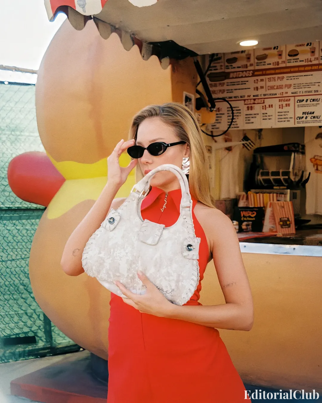 Ester Expósito in a red dress holding a textured Desigual handbag in front of a hot dog stand – Not a Doll campaign