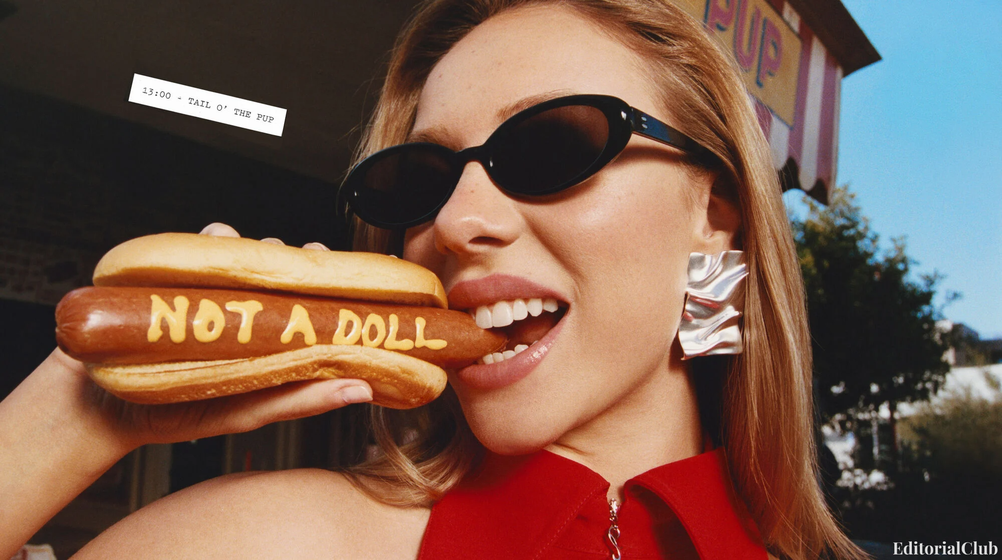 Ester Expósito bites into a hot dog with 'Not a Doll' written in mustard, wearing bold silver earrings and black sunglasses for Desigual’s 2025 campaign.