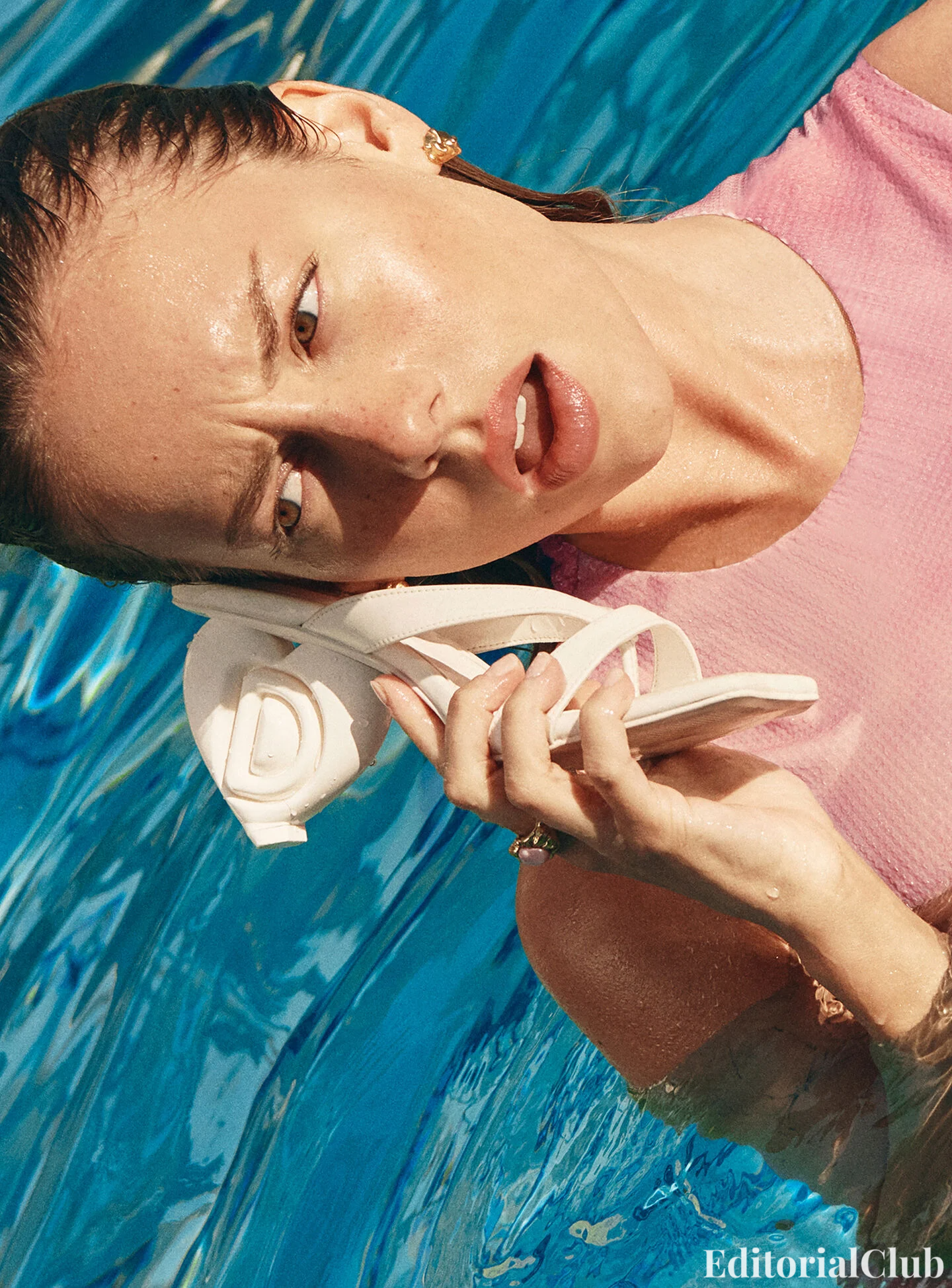 Ester Expósito playfully holds a white Desigual heel like a phone while relaxing in a pool, wearing a pink dress – campaign shoot